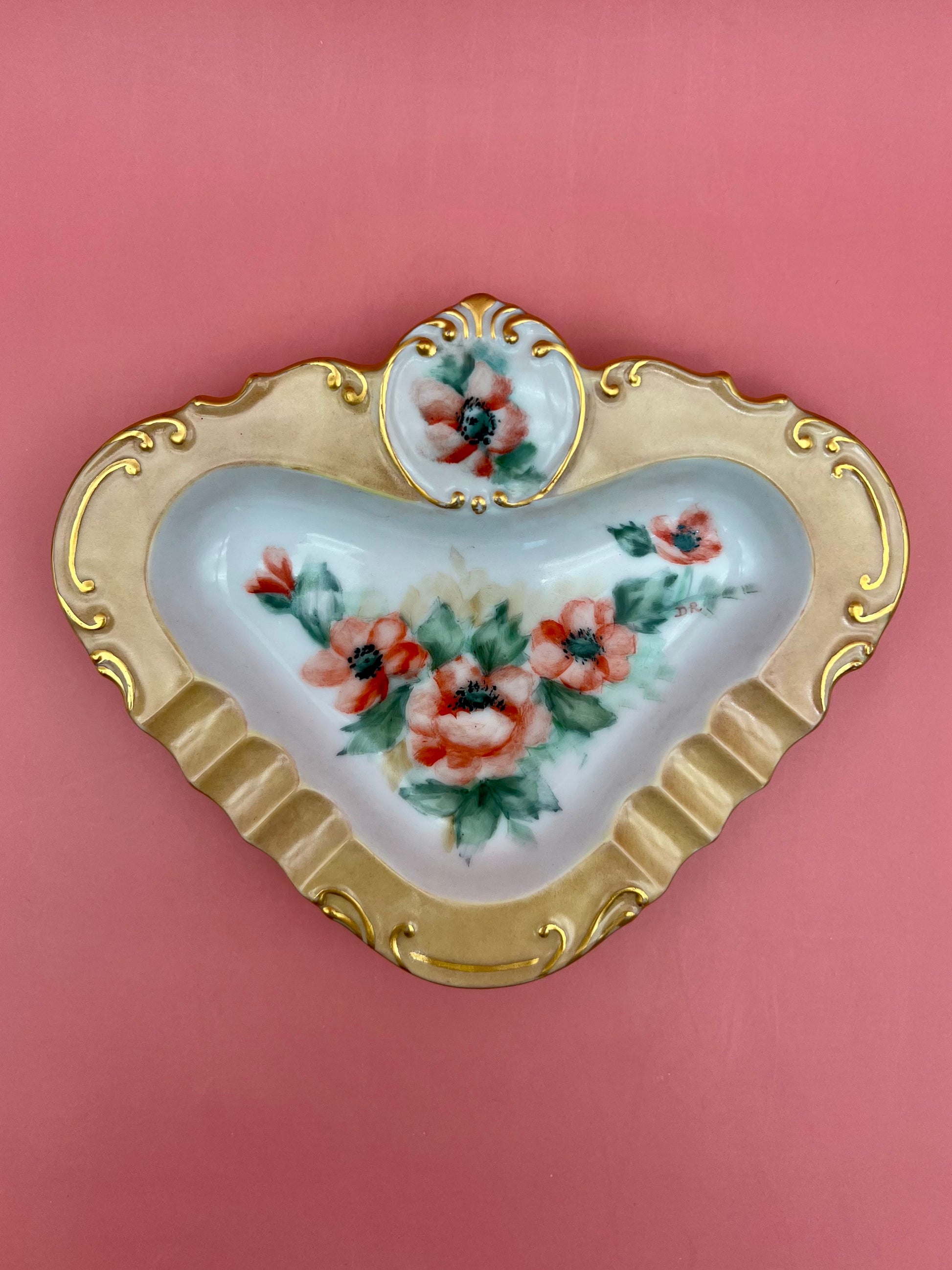 Straight-on view of the same triangular porcelain ashtray showing the gold scrollwork border and coral pink floral pattern, pink background.