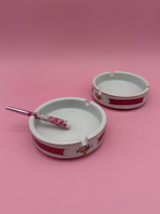 Two round white ceramic ashtrays with a pink floral border band, glass cigarette pick resting in one, pink background.
