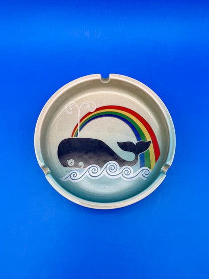 Round ceramic ashtray with a black whale swimming beneath a rainbow, blue background