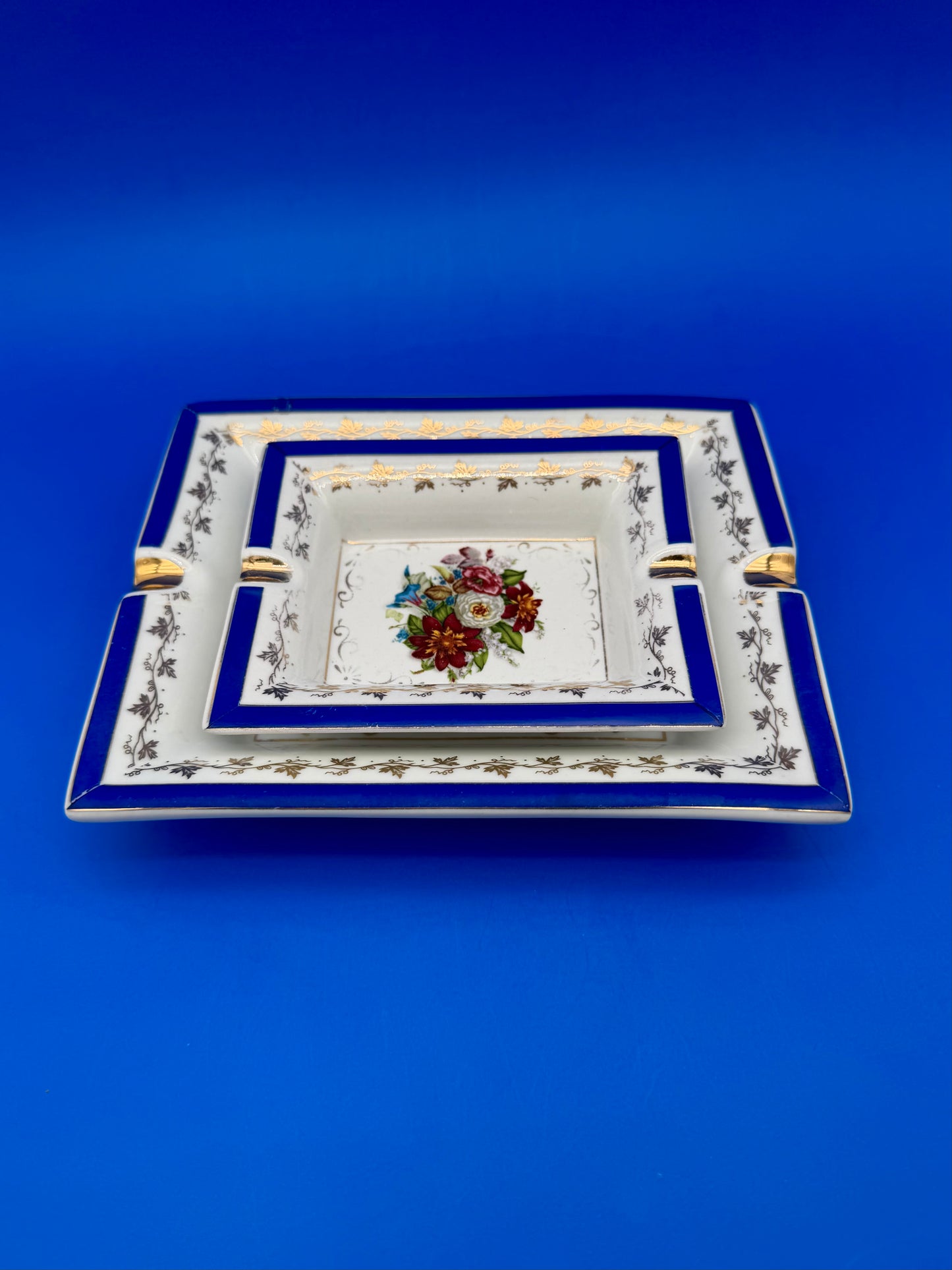 Rectangular white porcelain ashtray with cobalt blue border, gold vine trim, and a floral bouquet center, blue background