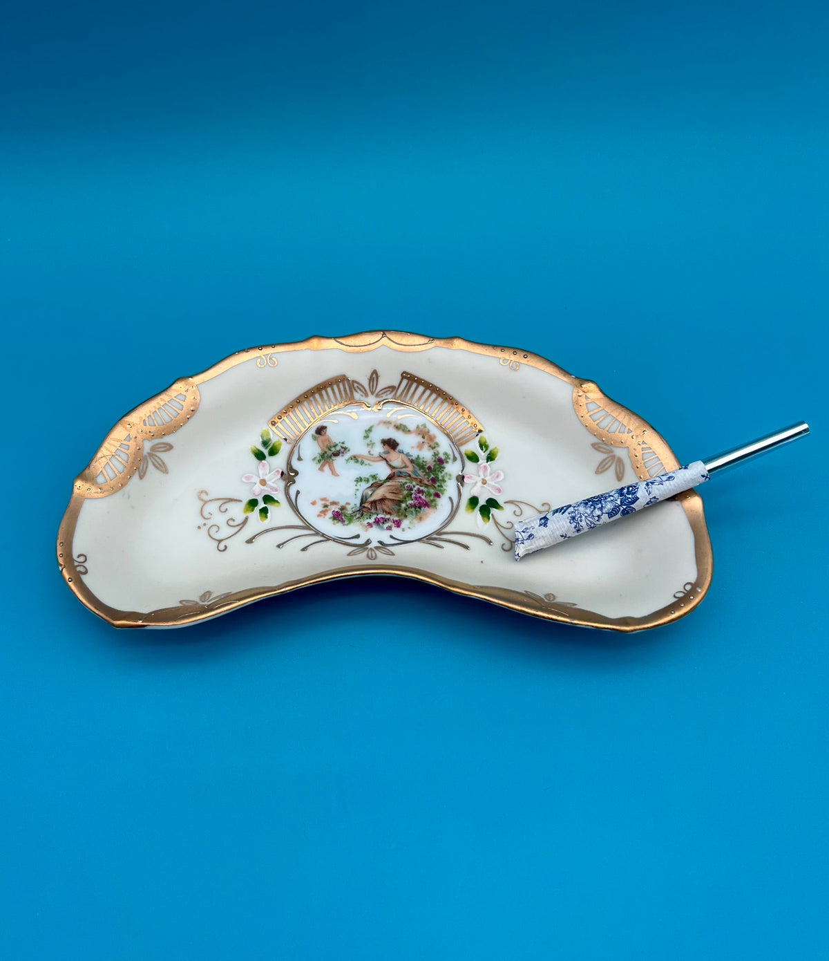 Crescent-shaped white porcelain ashtray with flowing gold trim framing a painted center medallion, glass cigarette pick resting across it, blue background.