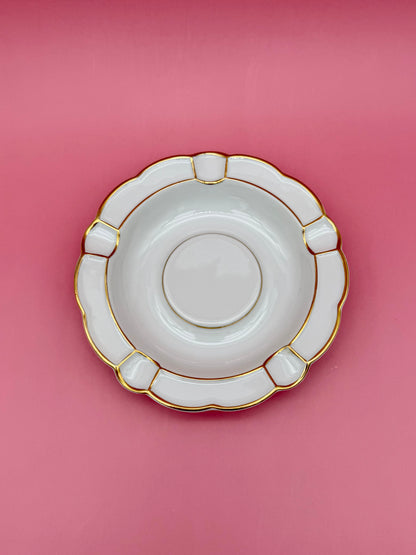 Same round white ashtray with paneled scalloped border and gold trim, shown without the cigarette pick, pink background