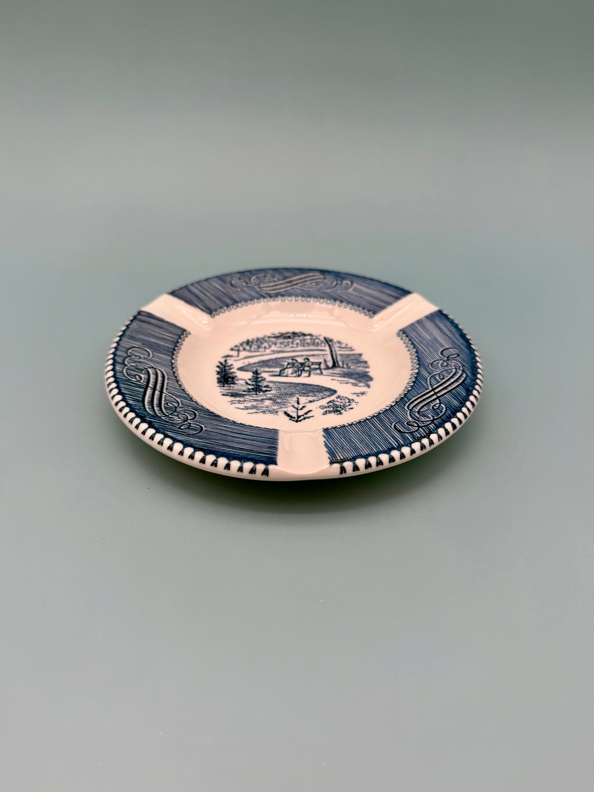 Round white ashtray with navy blue scalloped border and scenic landscape illustration, shown without the cigarette pick, gray background