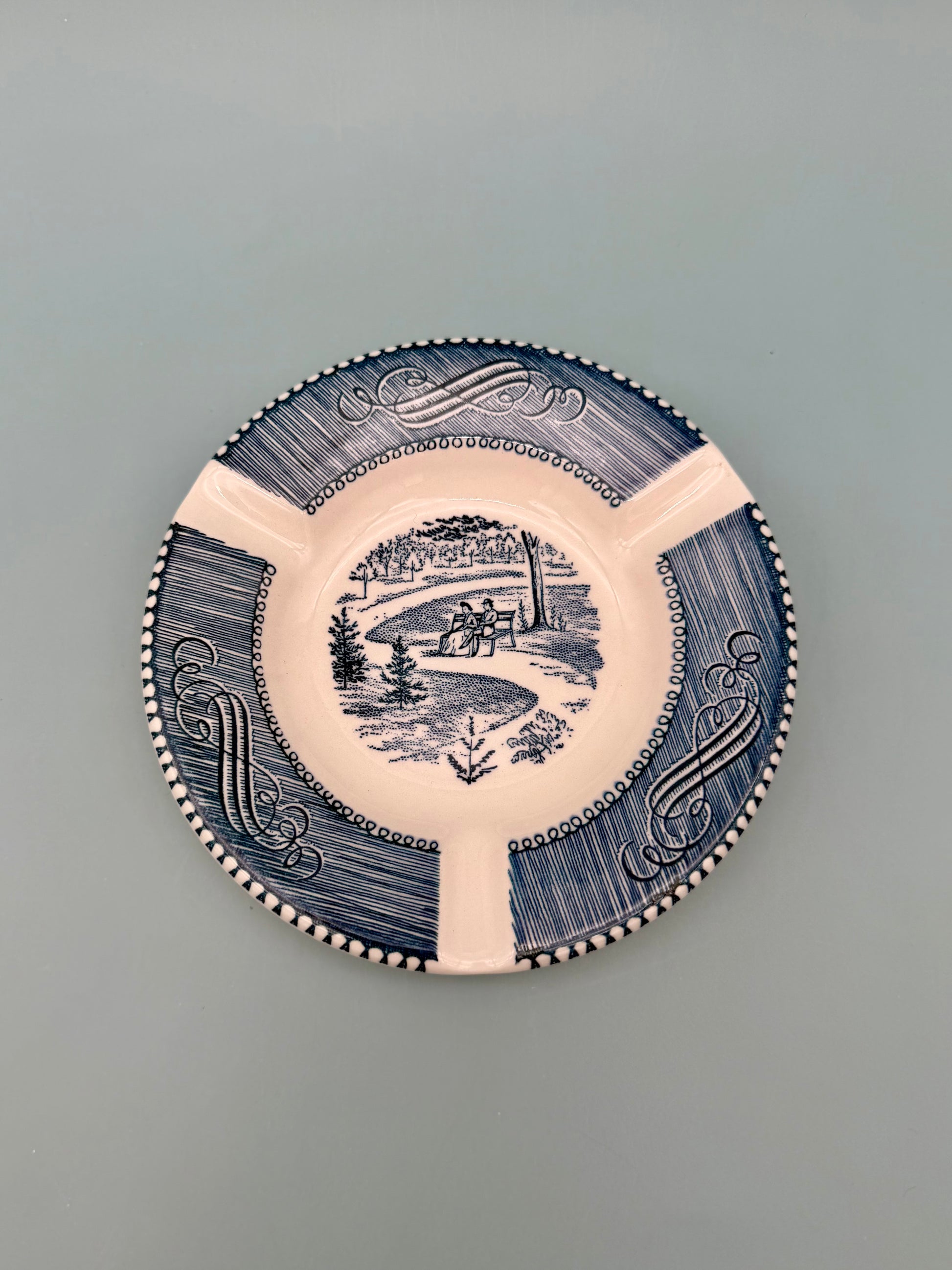 Round white ashtray with navy blue scalloped border and scenic landscape illustration, top-down close-up view, gray background
