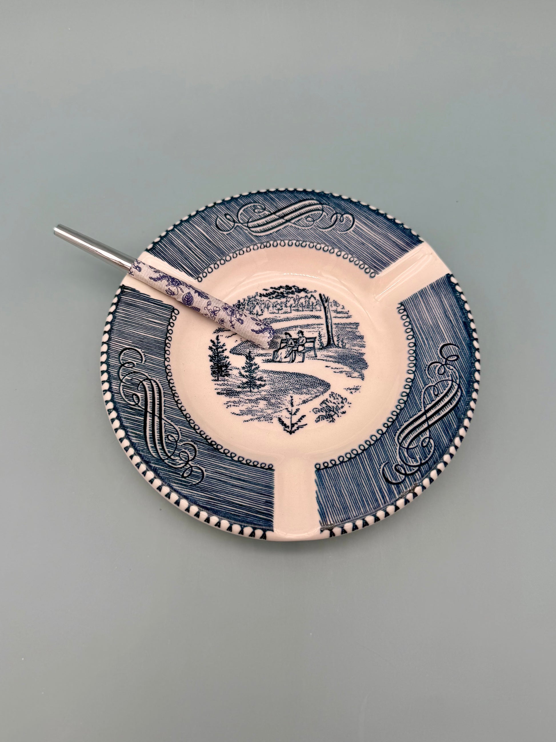 Round white ceramic ashtray with a navy blue scalloped border and a scenic landscape illustration at center, glass cigarette pick resting across it, gray background