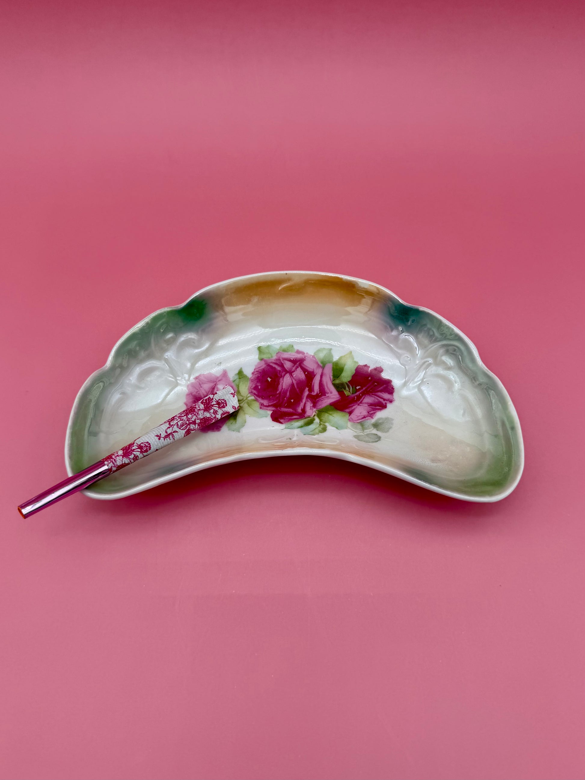 Crescent-shaped iridescent ceramic ashtray with a hand-painted pink rose design, glass cigarette pick resting across it, magenta background.