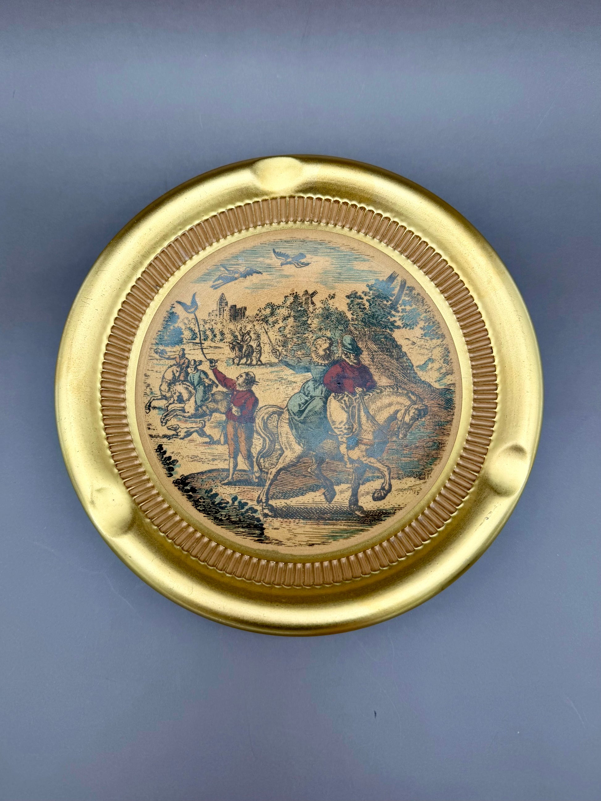 Round gold ceramic ashtray with a ribbed rim and a sepia-toned pastoral scene of figures and a horse in the center, gray-blue background.