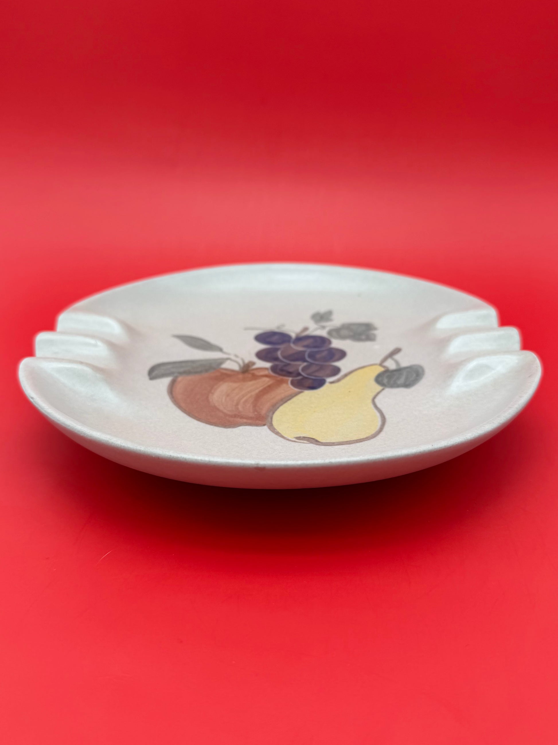Same round cream ashtray with the painted grape, apple, and pear cluster, shown without the cigarette pick, closer view, red background.