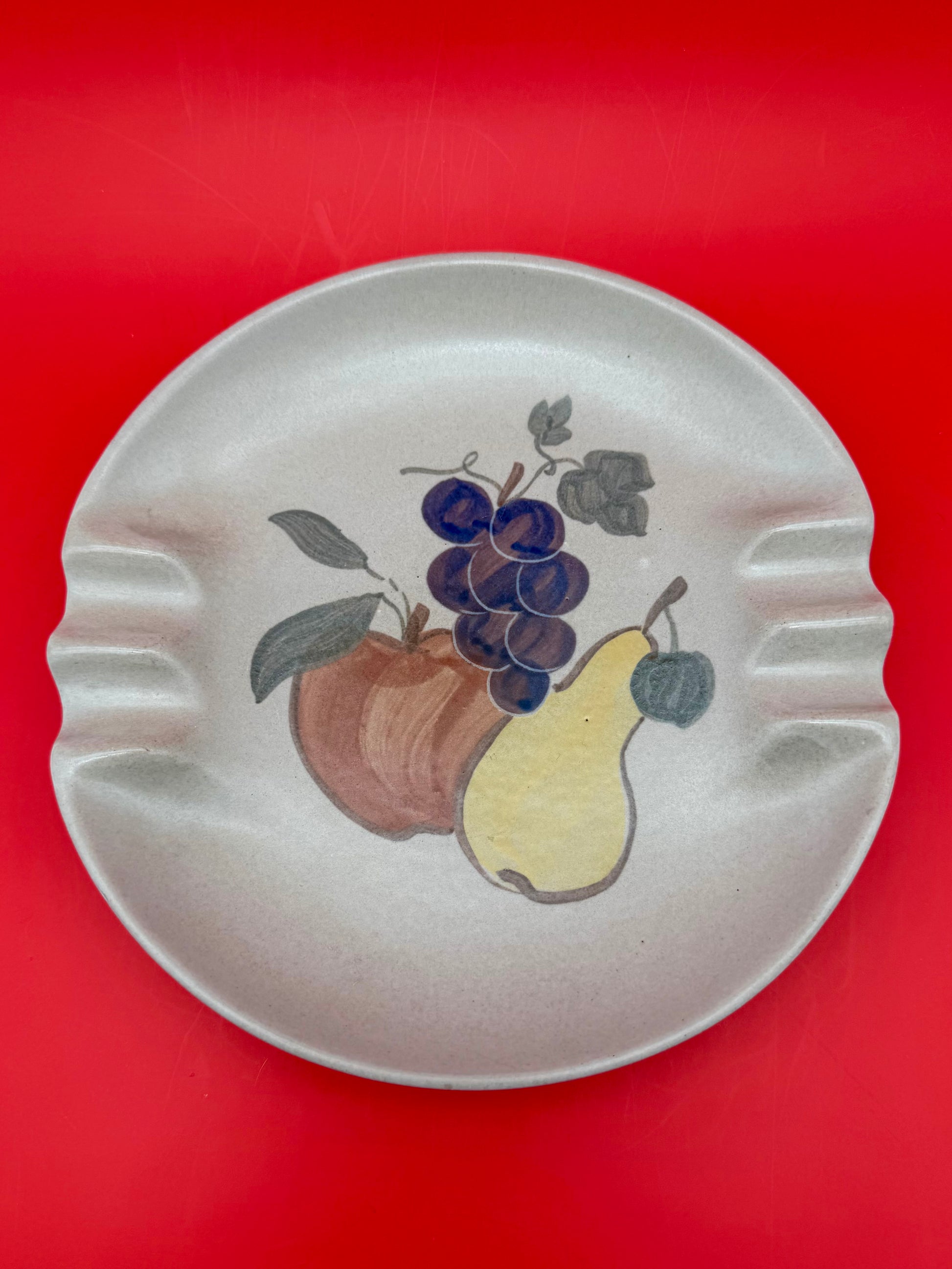 Round cream ceramic ashtray hand-painted with a cluster of grapes, an apple, and a pear, red background.
