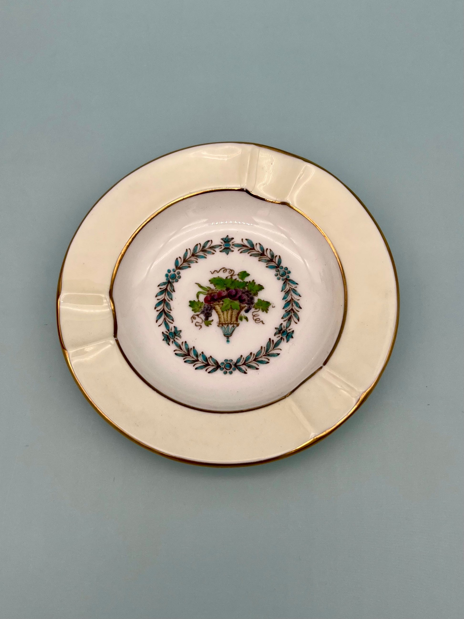 Round ceramic ashtray with a wide cream and gold rim framing a botanical basket motif in the center, sage background.