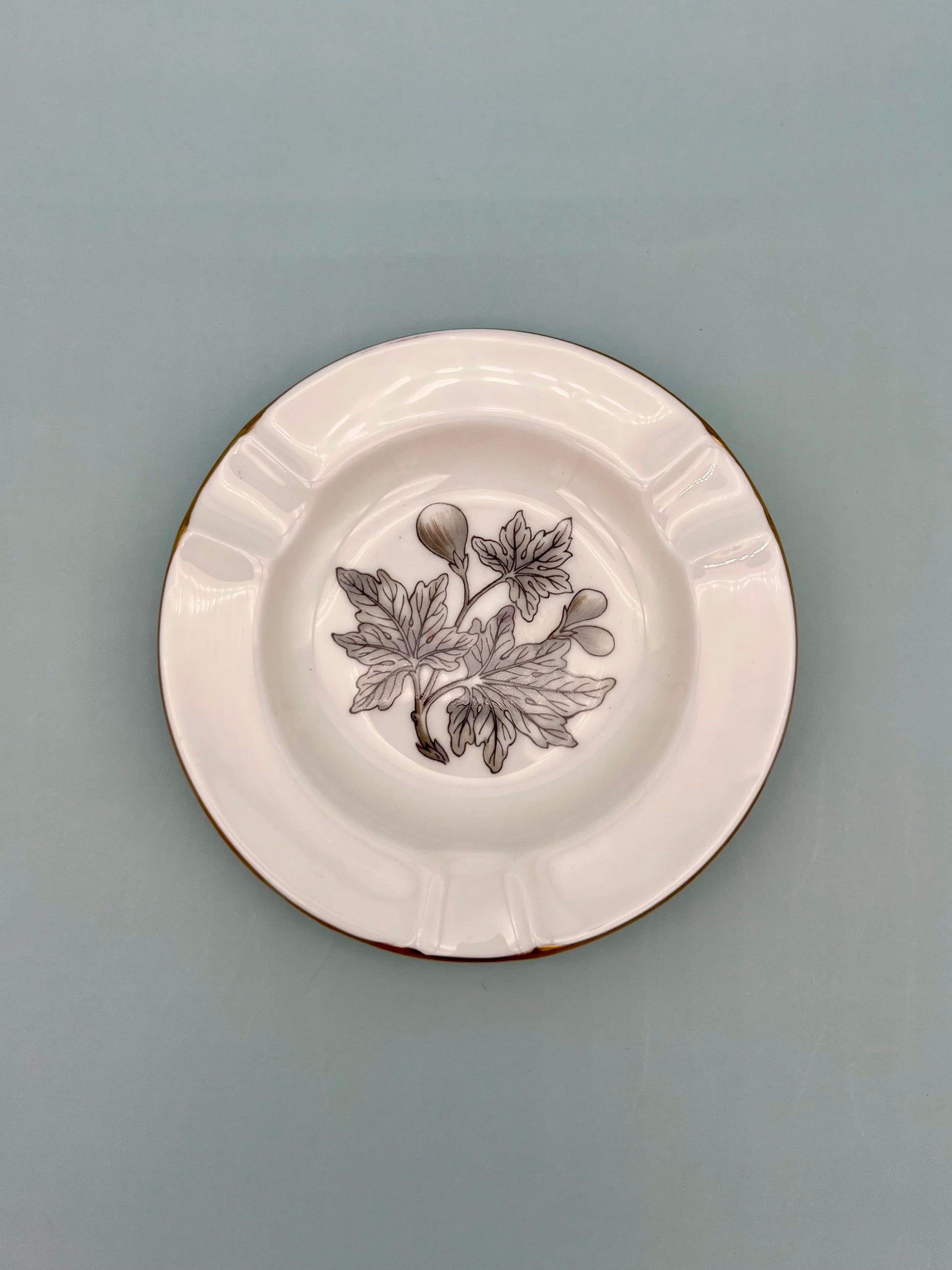 Round cream ceramic ashtray with a black-and-white botanical leaf illustration in the center and a scalloped rim, sage background.