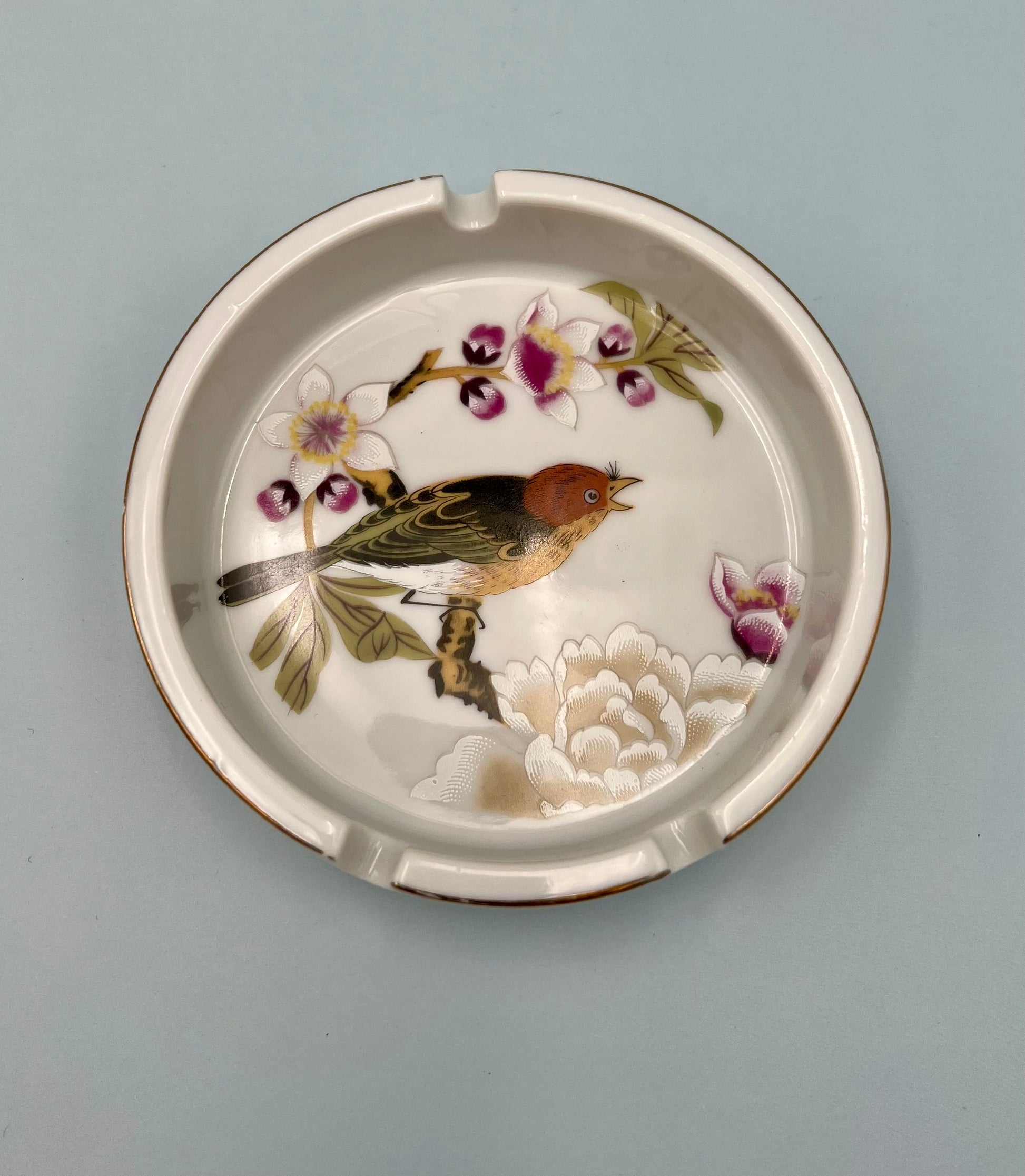 Round cream ceramic ashtray hand-painted with a bird perched among cherry blossoms, sage green background.