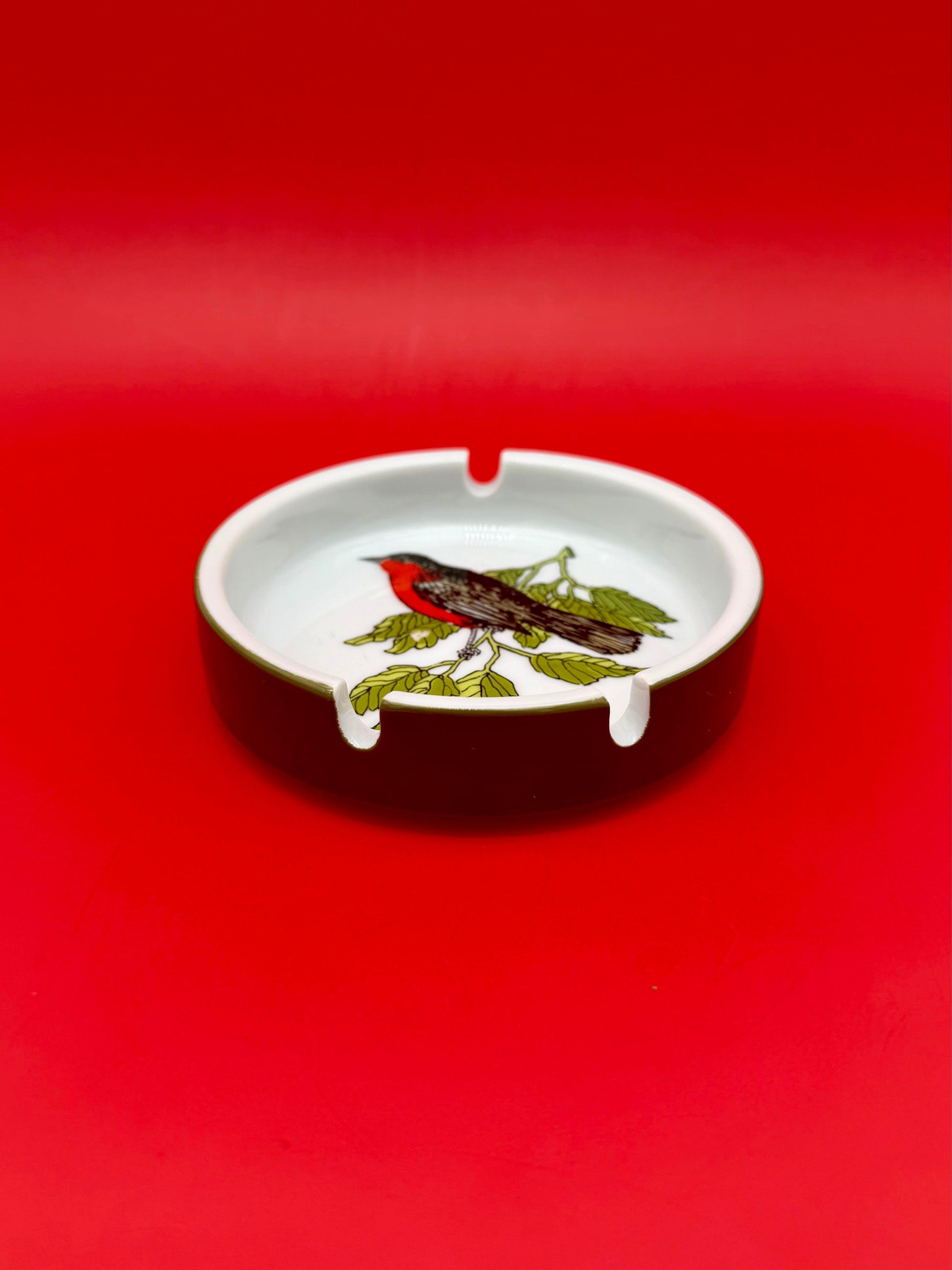 Low-angle view of a round white ashtray hand-painted with a reddish robin-like bird perched on a leafy branch, red background.