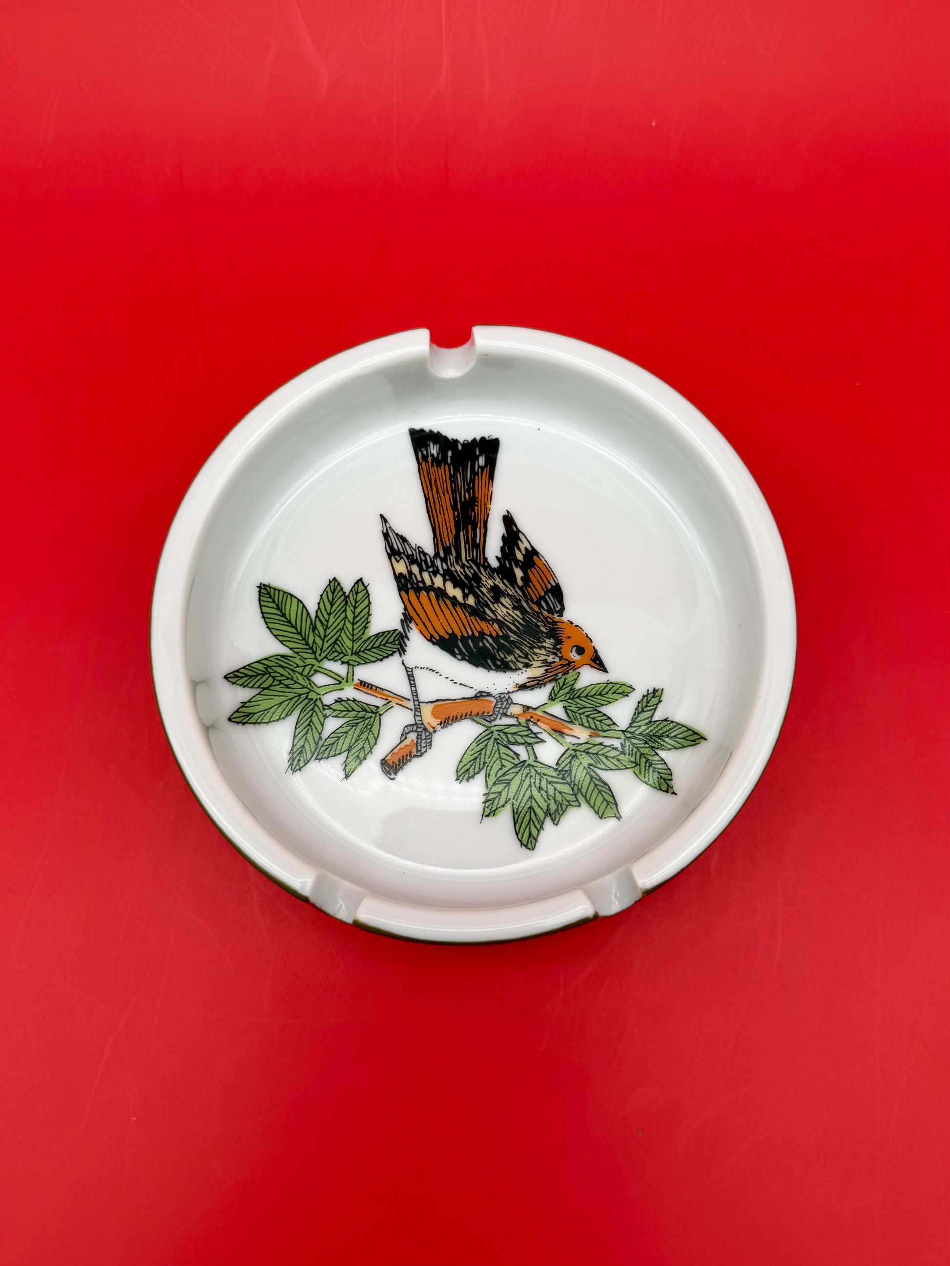 Top-down close-up of a round white ashtray hand-painted with an orange, black, and white bird perched on a leafy branch, red background.