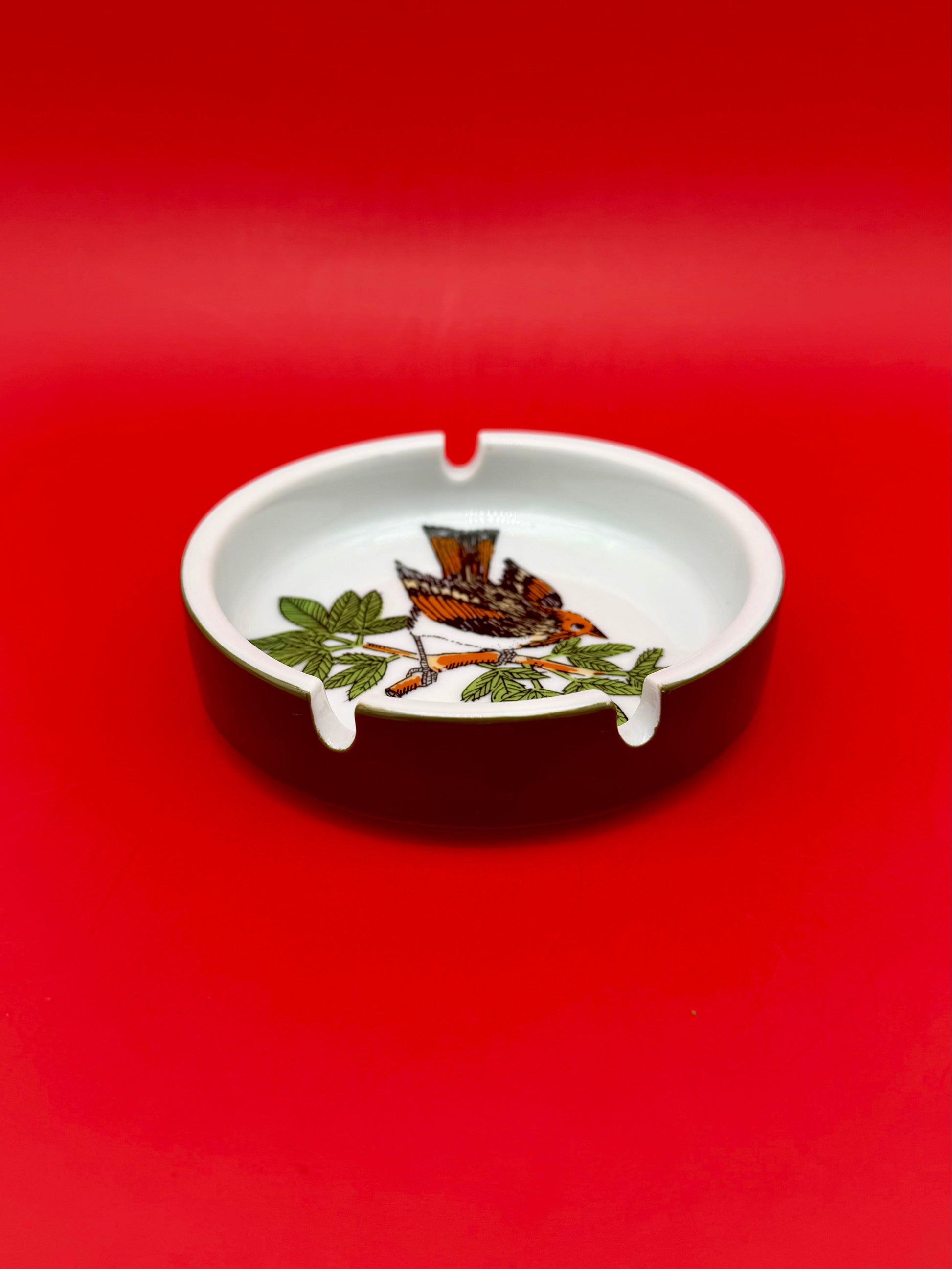 Low-angle view of the ashtray with the orange, black, and white bird perched on a leafy branch, red background.
