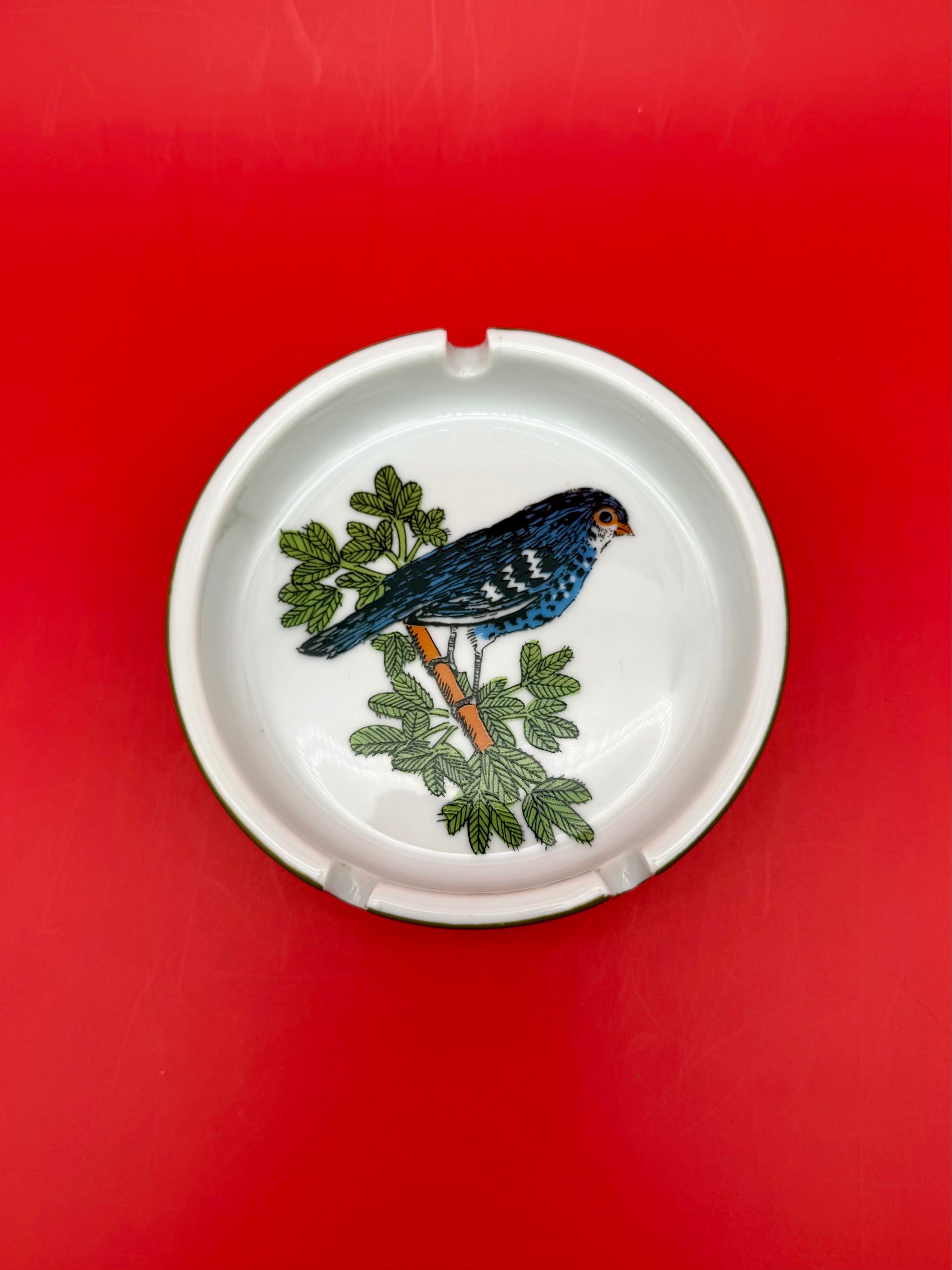 Top-down close-up of a round white ashtray hand-painted with a blue-gray bird perched on a leafy branch, red background.