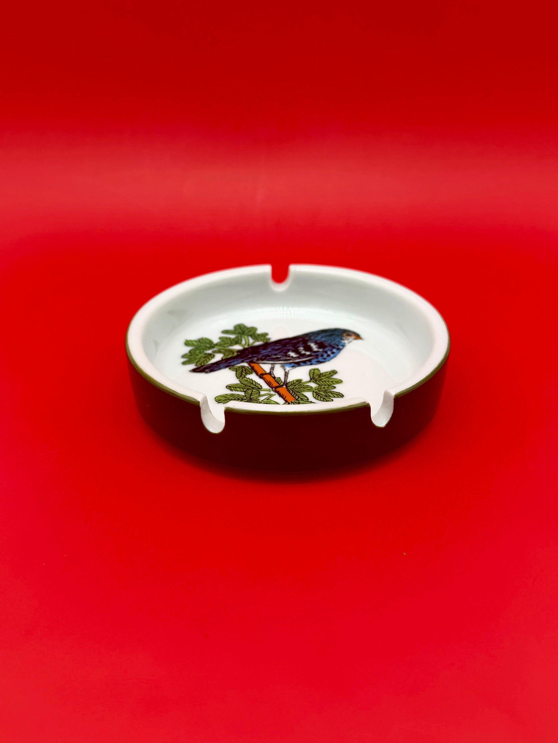 Low-angle view of a round white ashtray hand-painted with a bird in warm brown and orange tones perched on a leafy branch, red background.