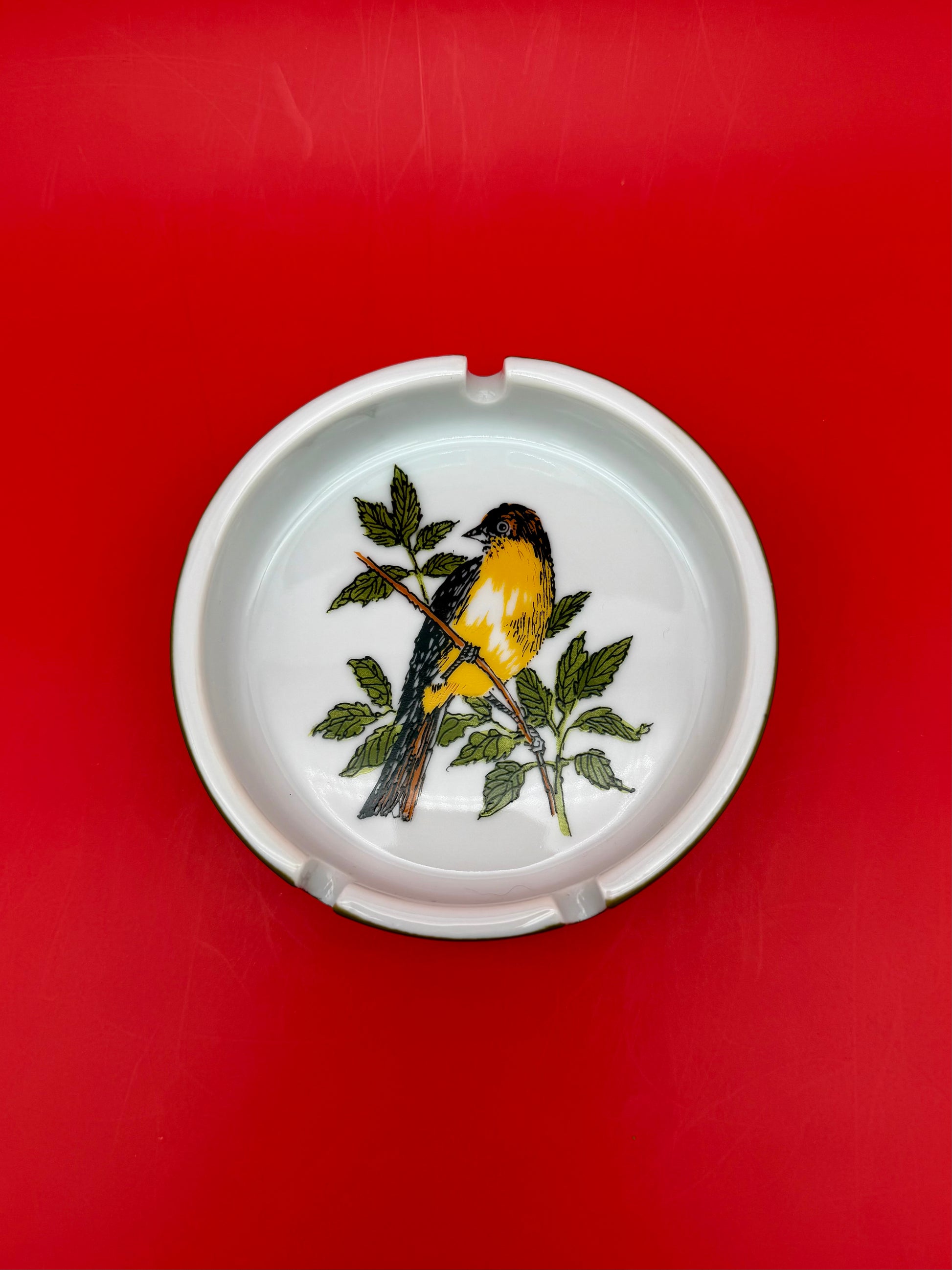 Top-down close-up of a round white ashtray hand-painted with a yellow and black bird perched on a leafy branch, red background.