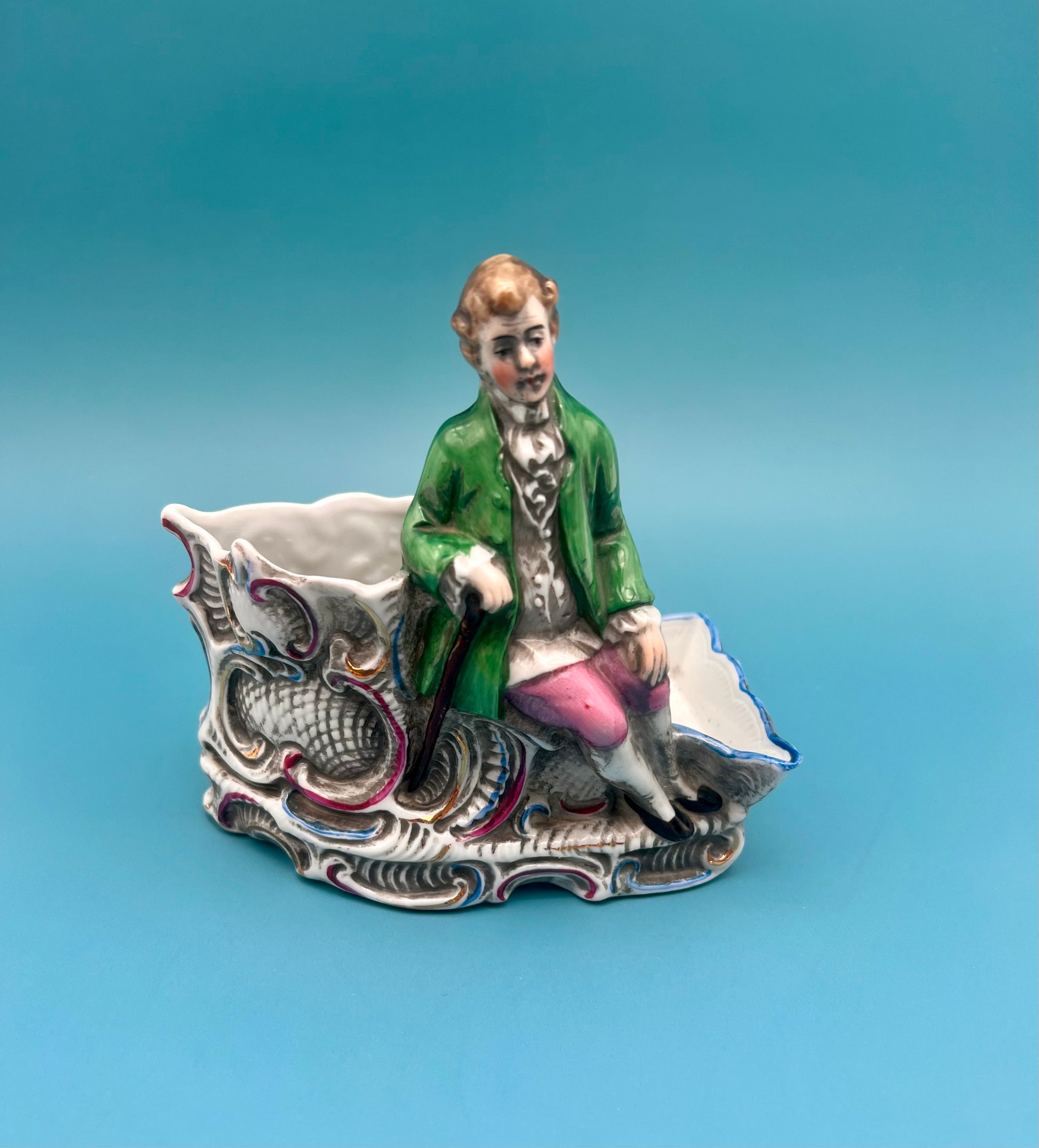 Closer view of the Ludwig Set gentleman figurine in a green coat and pink breeches seated on the scrollwork base, teal background.