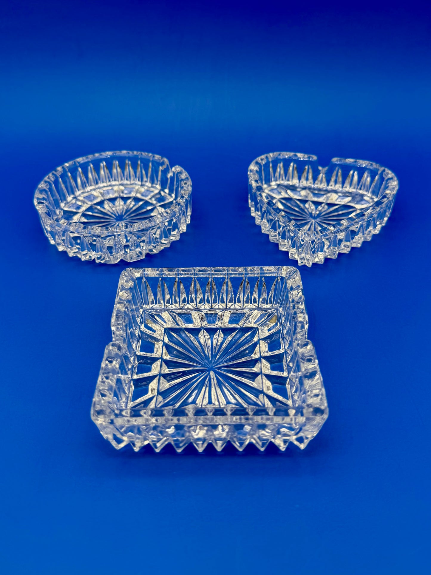Round, heart-shaped, and square faceted crystal ashtrays shown from a different angle, blue background