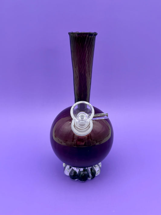 Purple glass decanter-shaped water pipe with a tall fluted trumpet neck and round bulb base on a scalloped foot, glass bowl piece attached at the front, purple background.