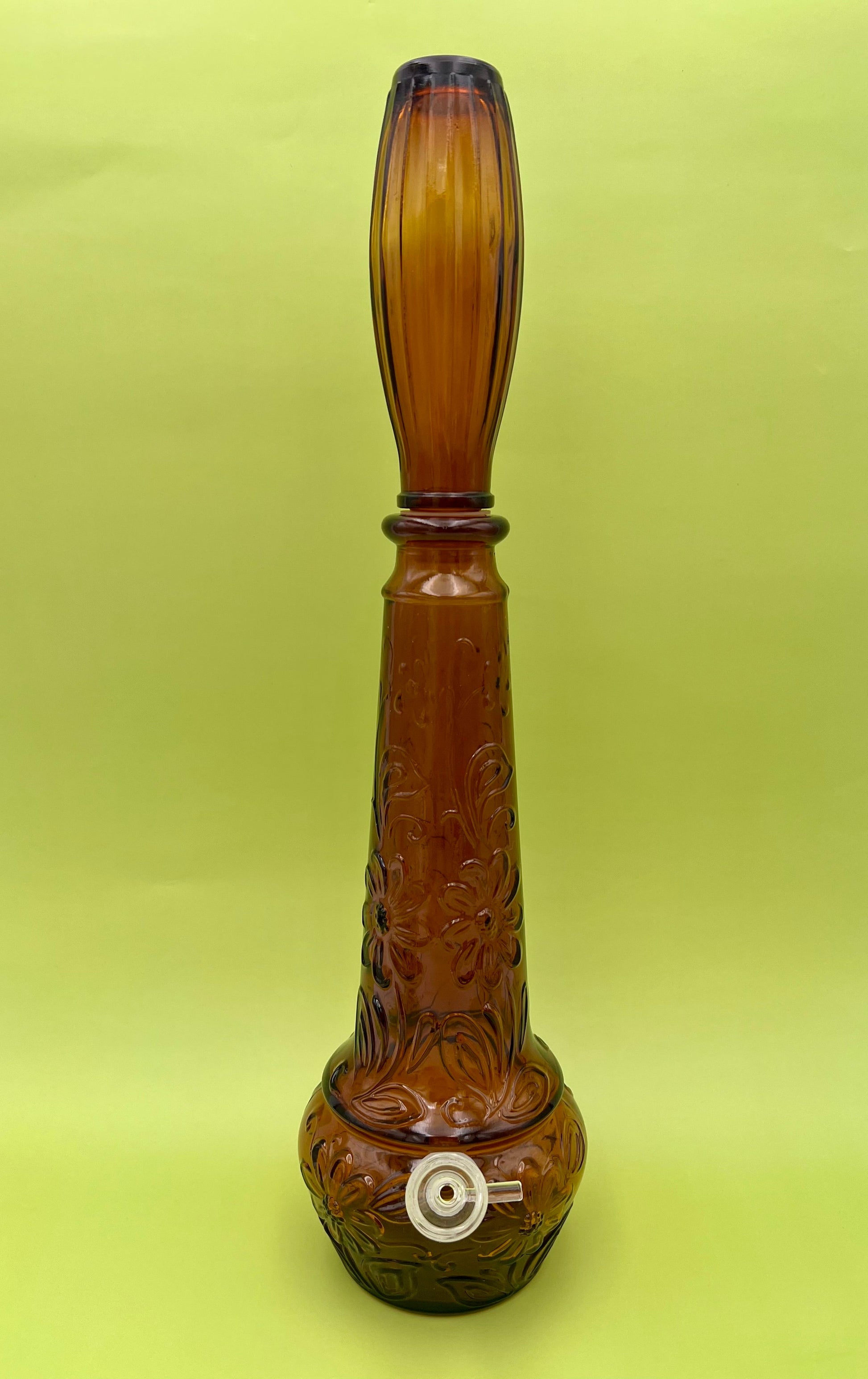 Front view of an amber glass genie-bottle decanter water pipe with embossed daisy pattern and ribbed stopper, glass bowl piece on the side, chartreuse background.