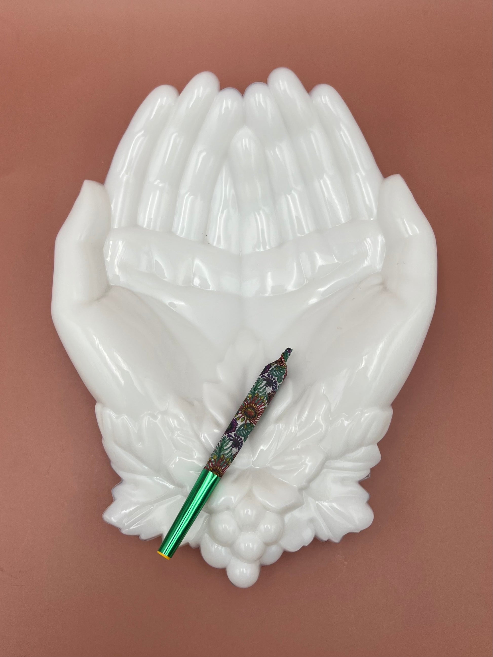 White porcelain ashtray shaped like two cupped hands, with a glass cigarette pick resting across the palms, mauve background.