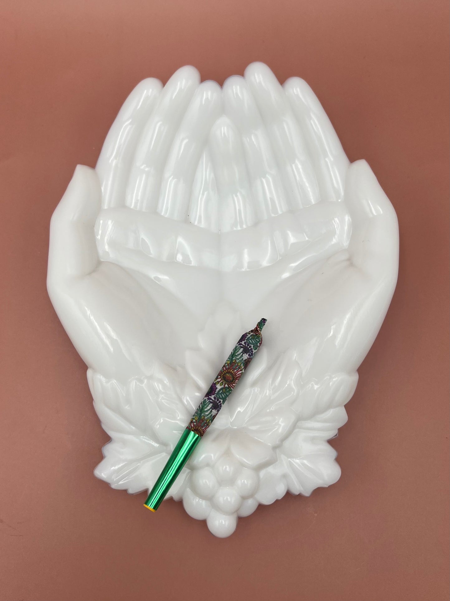 White porcelain ashtray shaped like two cupped hands, with a glass cigarette pick resting across the palms, mauve background.