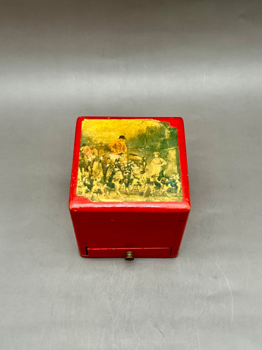Small red lacquered square trinket box with a hand-painted rural landscape scene in yellow and green tones on the lid, gray background.