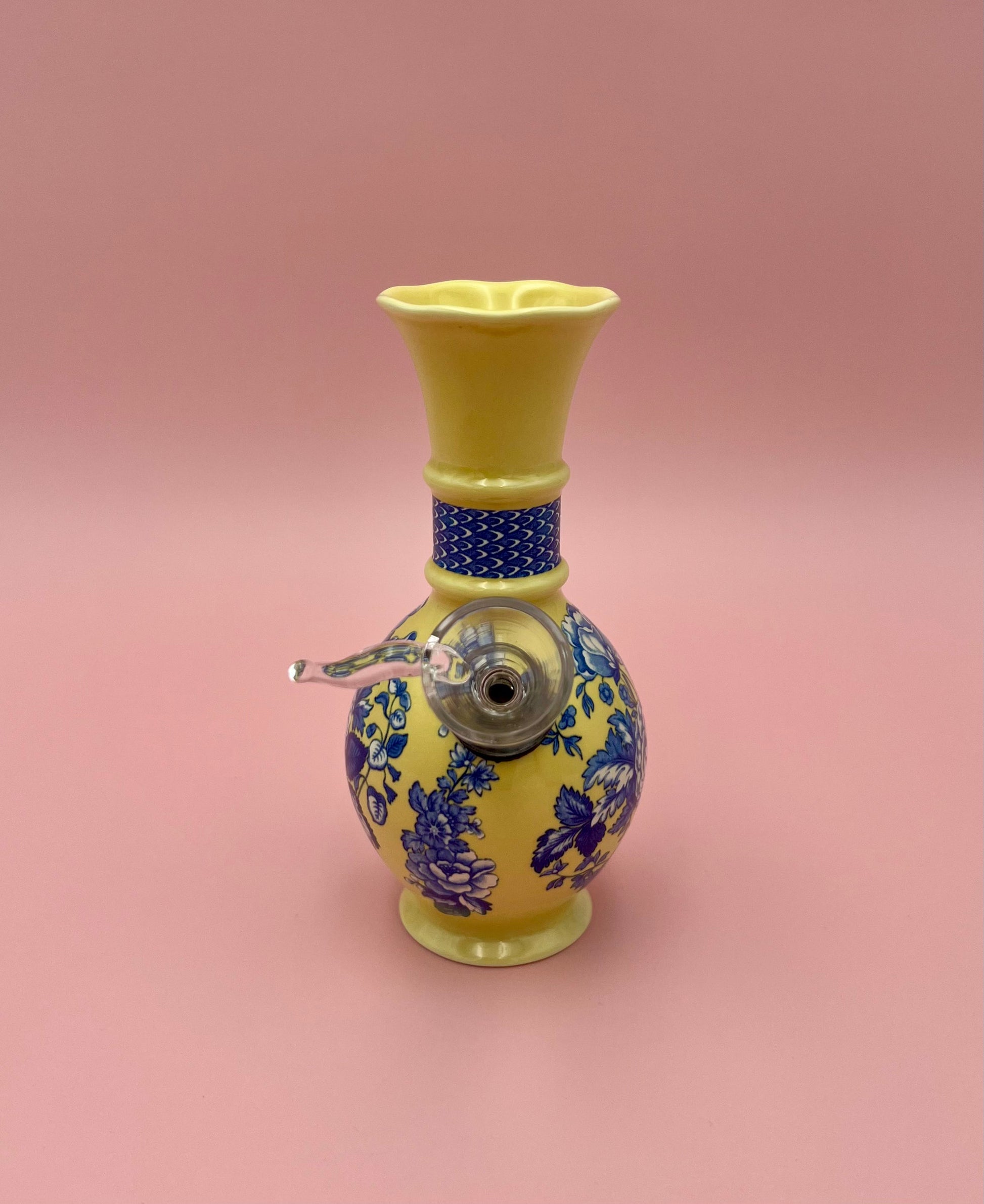 Front view of a yellow porcelain bud vase water pipe with a blue floral band and metallic bowl piece attached at the side, pink background.