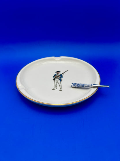 Same white ashtray with a painted soldier figure, now with a glass cigarette pick resting across it, blue background.