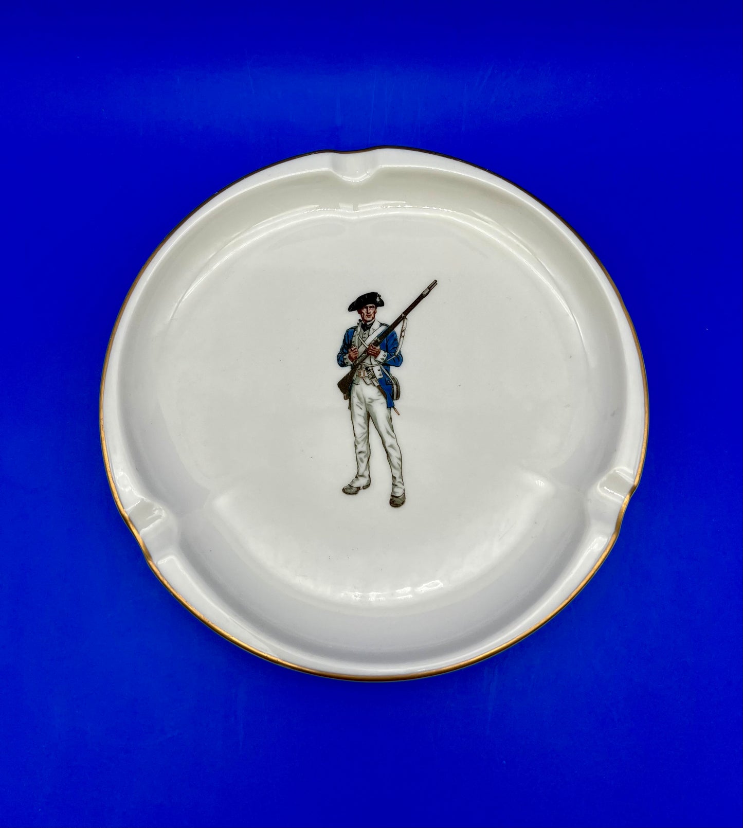 Round white ceramic ashtray hand-painted with a soldier figure holding a rifle, blue background.