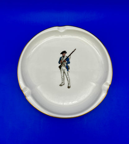 Round white ceramic ashtray hand-painted with a soldier figure holding a rifle, blue background.
