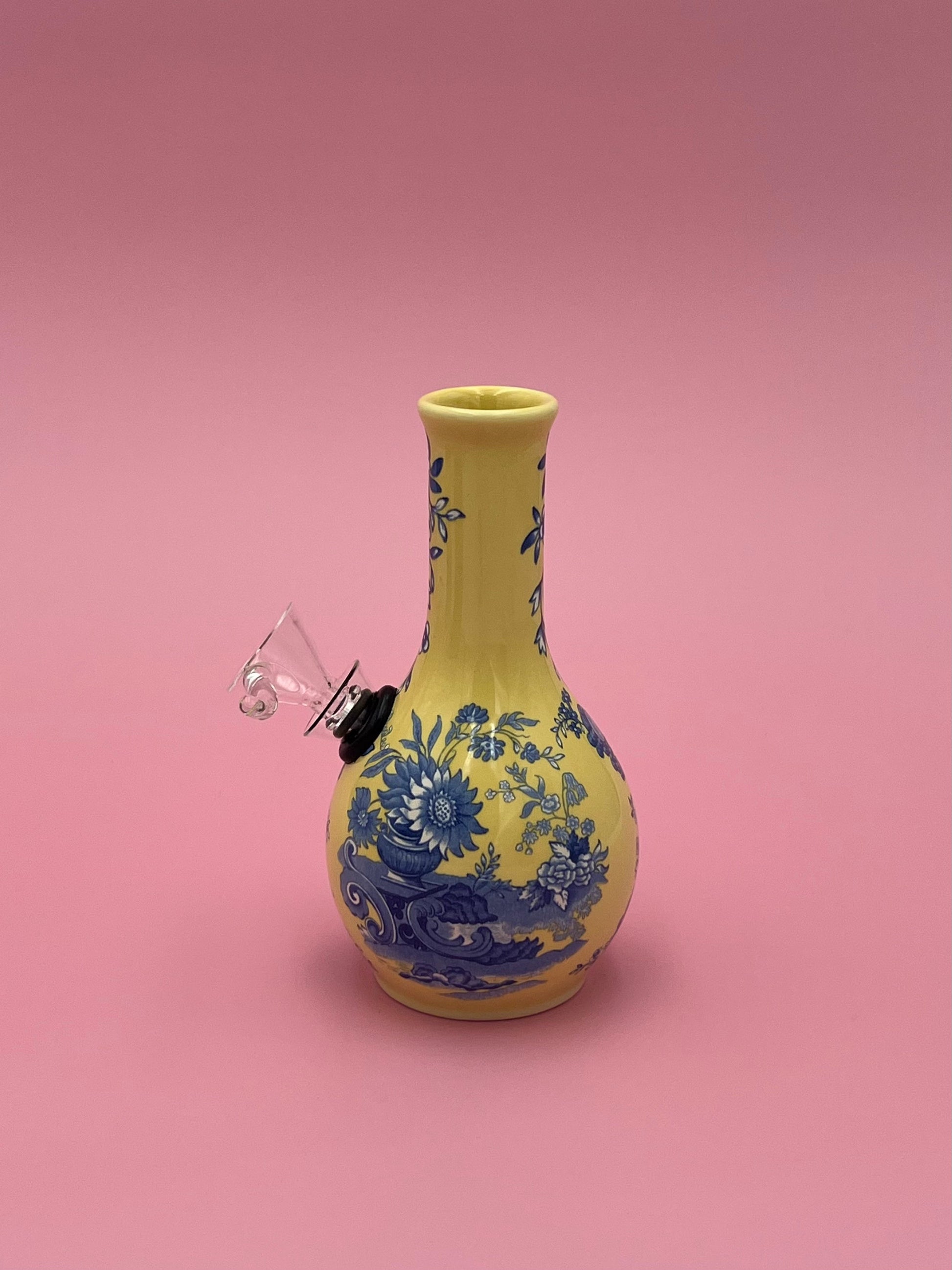 Front view of a butter-yellow porcelain bud vase water pipe with a cobalt blue floral toile pattern, glass bowl piece on the side, pink background.