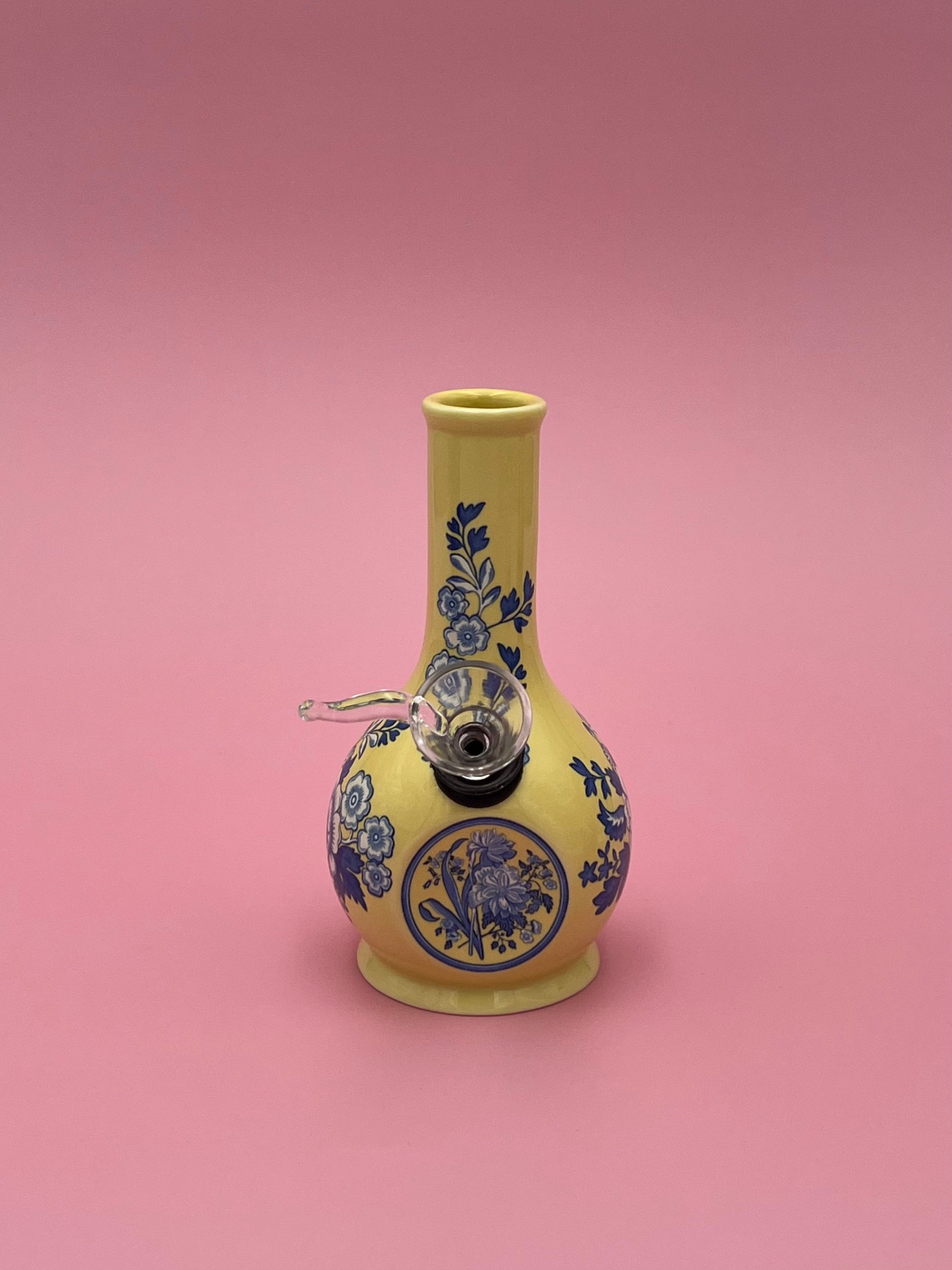 Front view of a white porcelain bud vase water pipe with a cobalt blue floral toile pattern and round medallion, glass bowl piece on the side, warm-toned pink background.