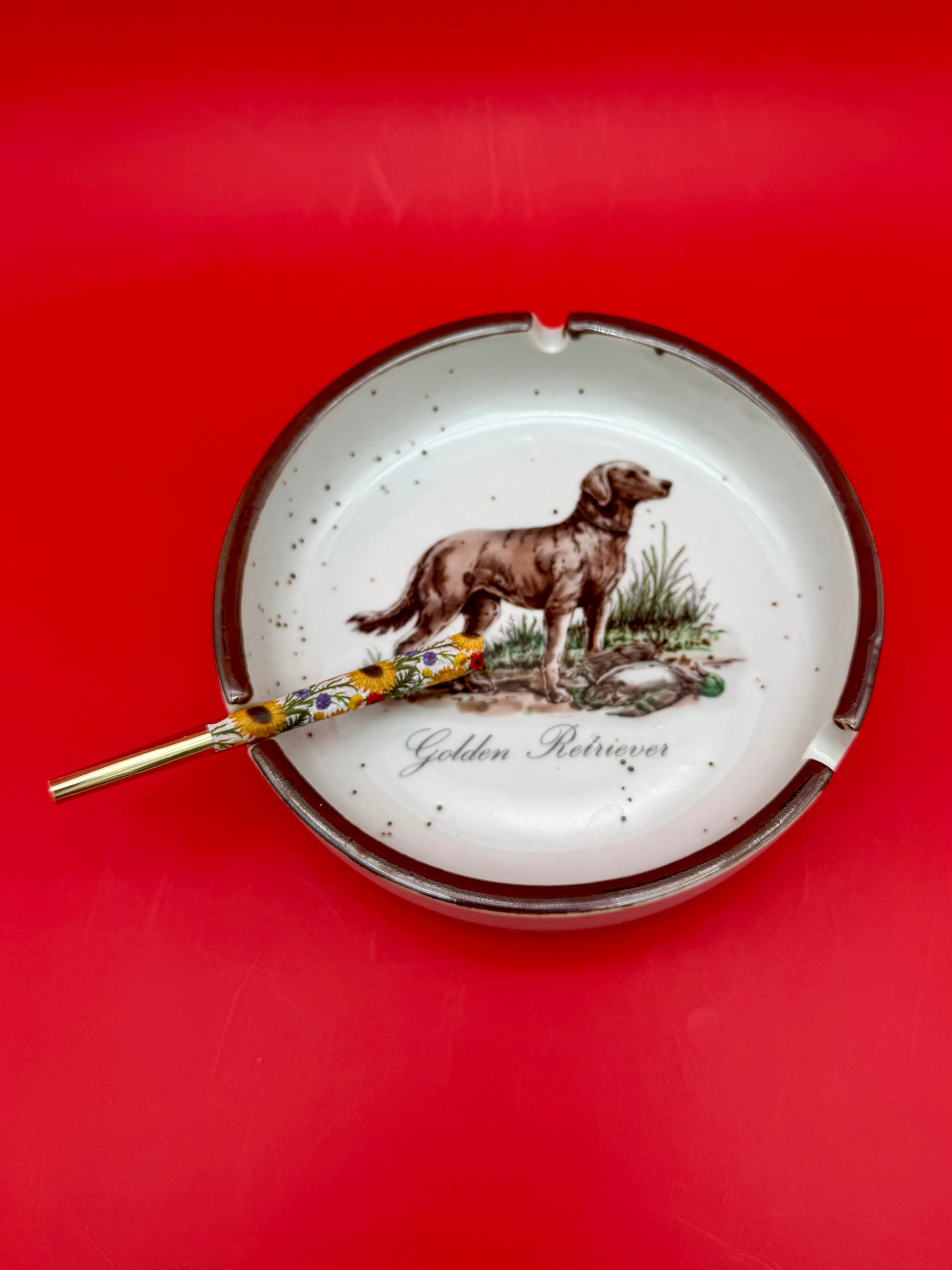 Oval white ceramic ashtray with a burgundy rim, hand-painted with a golden retriever dog and text, glass cigarette pick resting across it, red background.