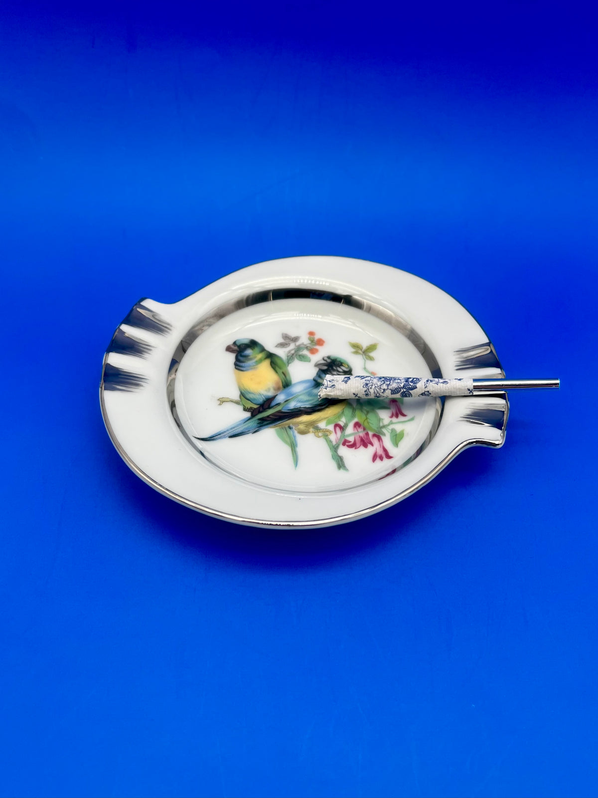 Same round white ashtray with a painted bluebird among blossoms, now with a glass cigarette pick with a floral handle resting across it, blue background.