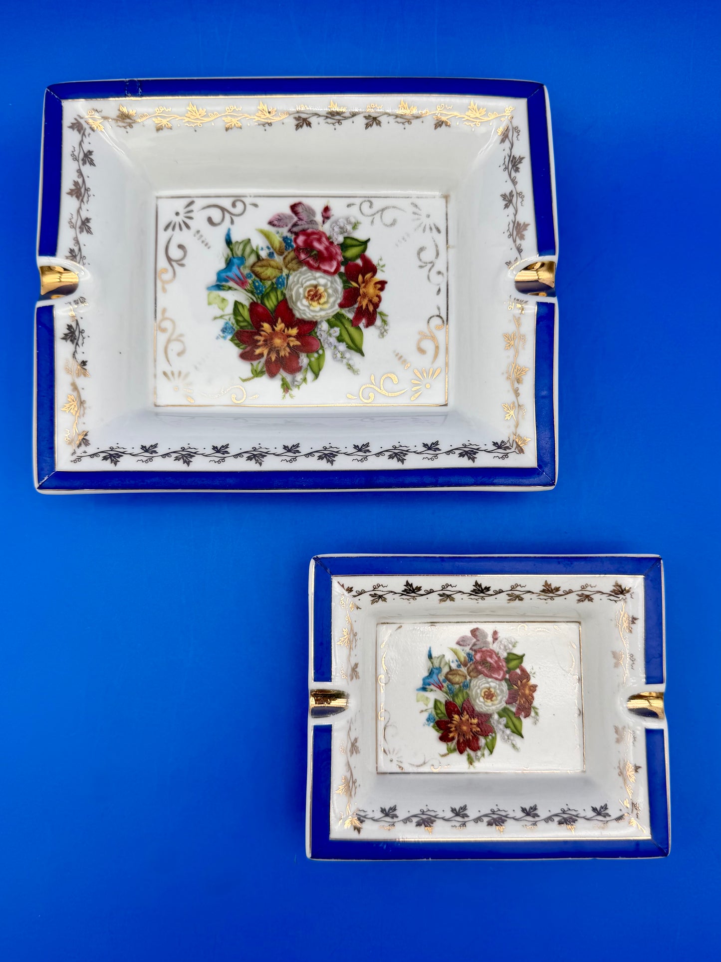 Two coordinating rectangular floral ashtrays in different sizes with cobalt blue borders and gold trim, blue background