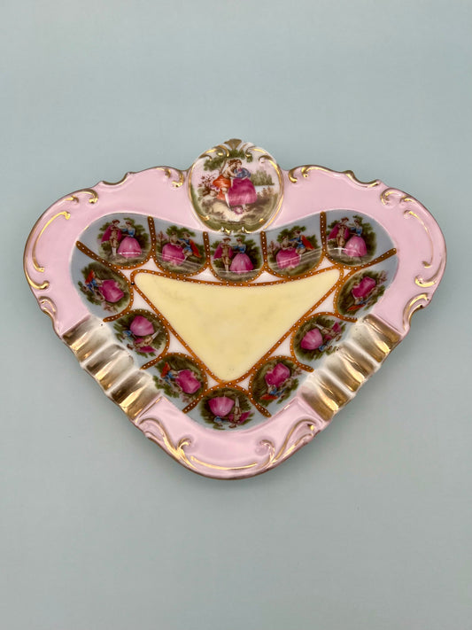 Heart-shaped pink and white porcelain tray with a scalloped gilt edge and a painted courting-couple medallion at the top, gray-blue background.