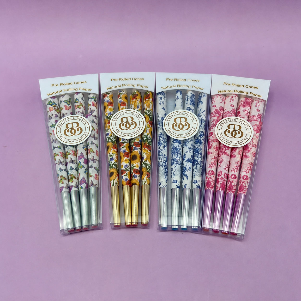Four packaged sets of floral-patterned pre-rolled cones with gold foil tips standing upright, purple background