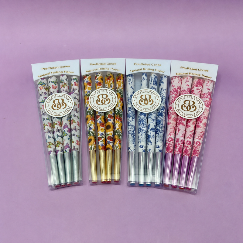 Four packaged sets of floral-patterned pre-rolled cones with gold foil tips standing upright, purple background