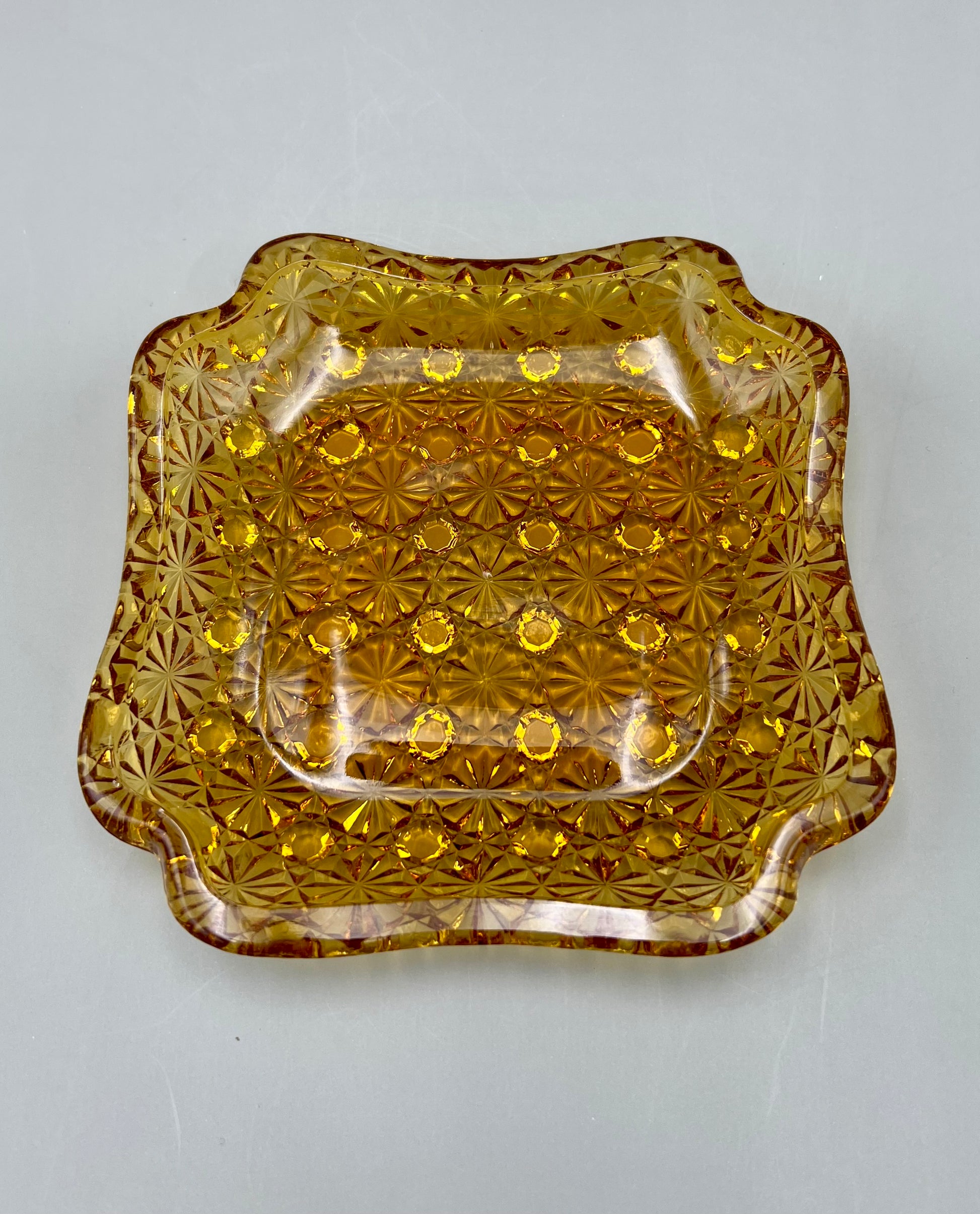 Amber pressed-glass ashtray with an embossed daisy and button pattern and scalloped edges, gray background