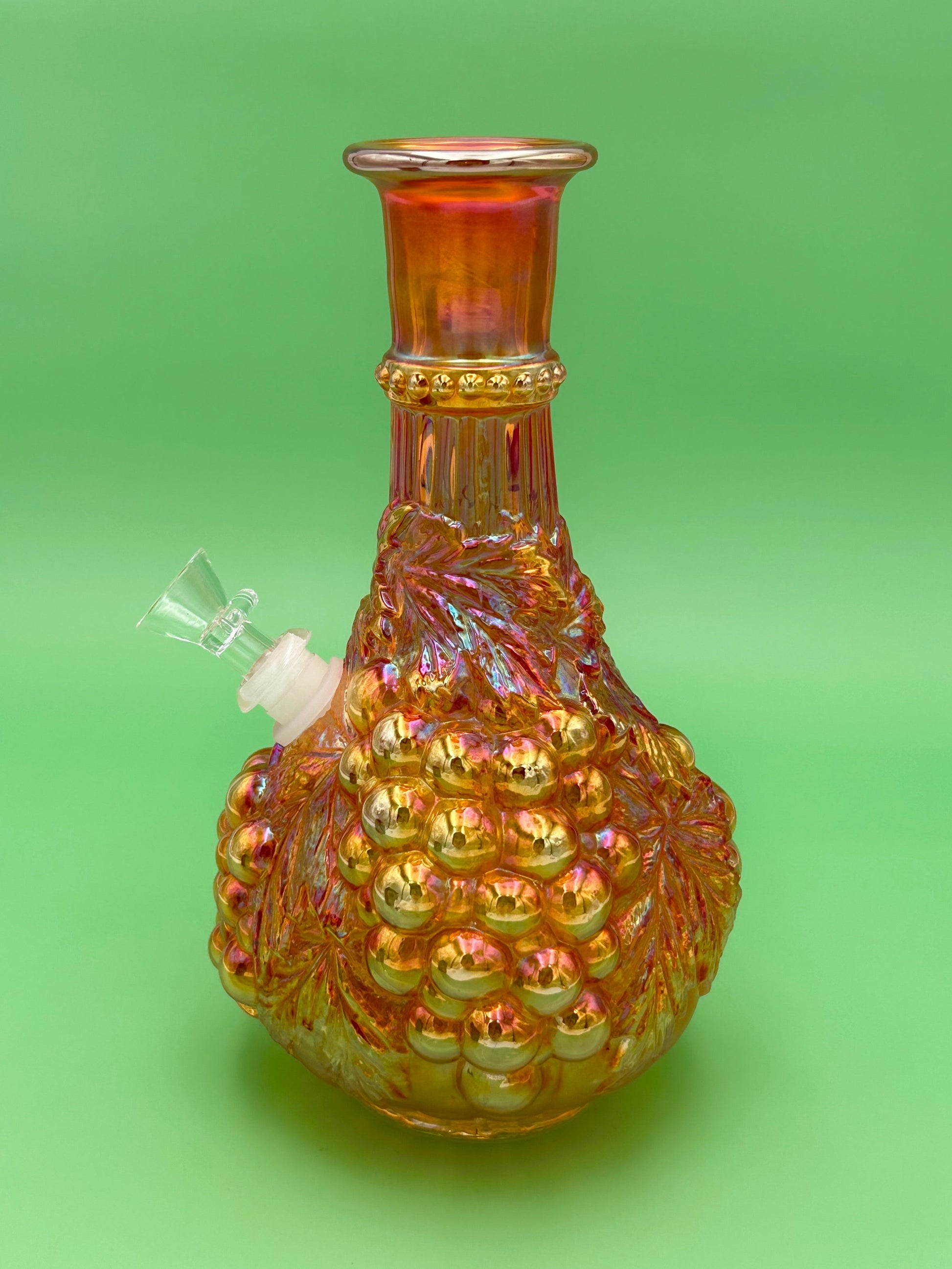 Marigold iridescent carnival glass decanter with an embossed grape and leaf pattern, white ceramic mouthpiece attached at the side, green background