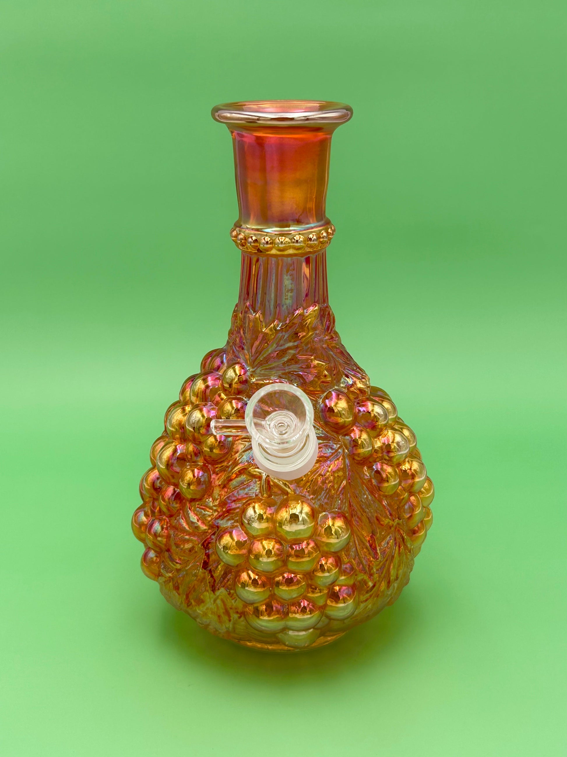 Front view of an iridescent marigold carnival glass decanter water pipe with an embossed grape pattern, glass bowl piece on the side, green background.