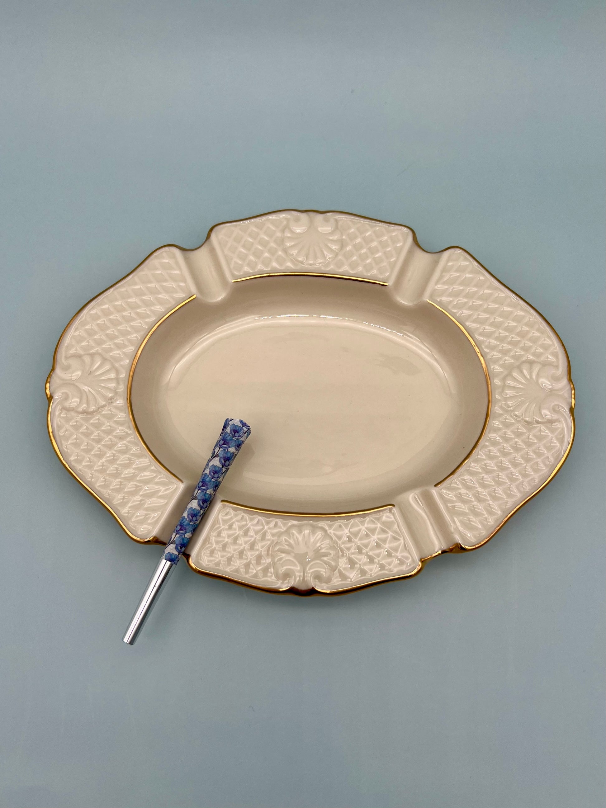 Oval cream ceramic ashtray with an embossed textured rim and gold trim, glass cigarette pick resting across it, blue-gray background.