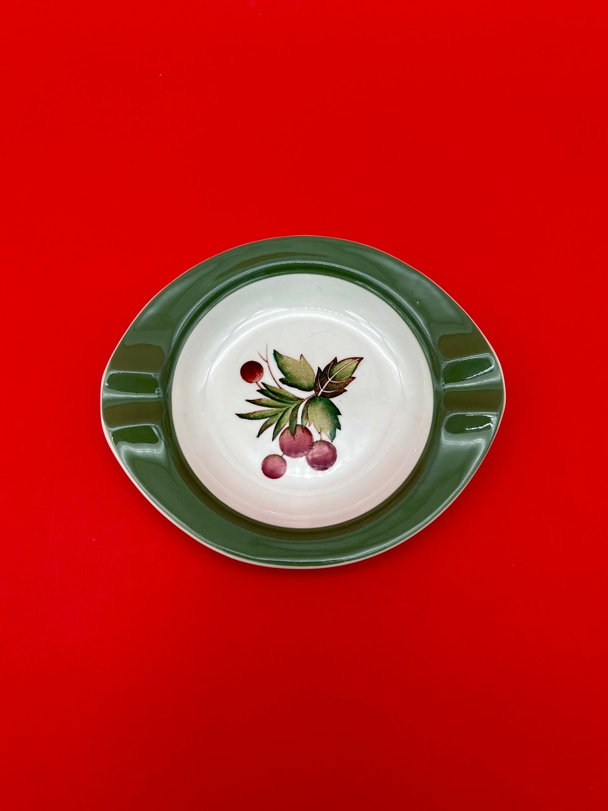 Same oval green-rimmed ashtray with the painted cherry branch design, shown as a closer top-down view, red background.