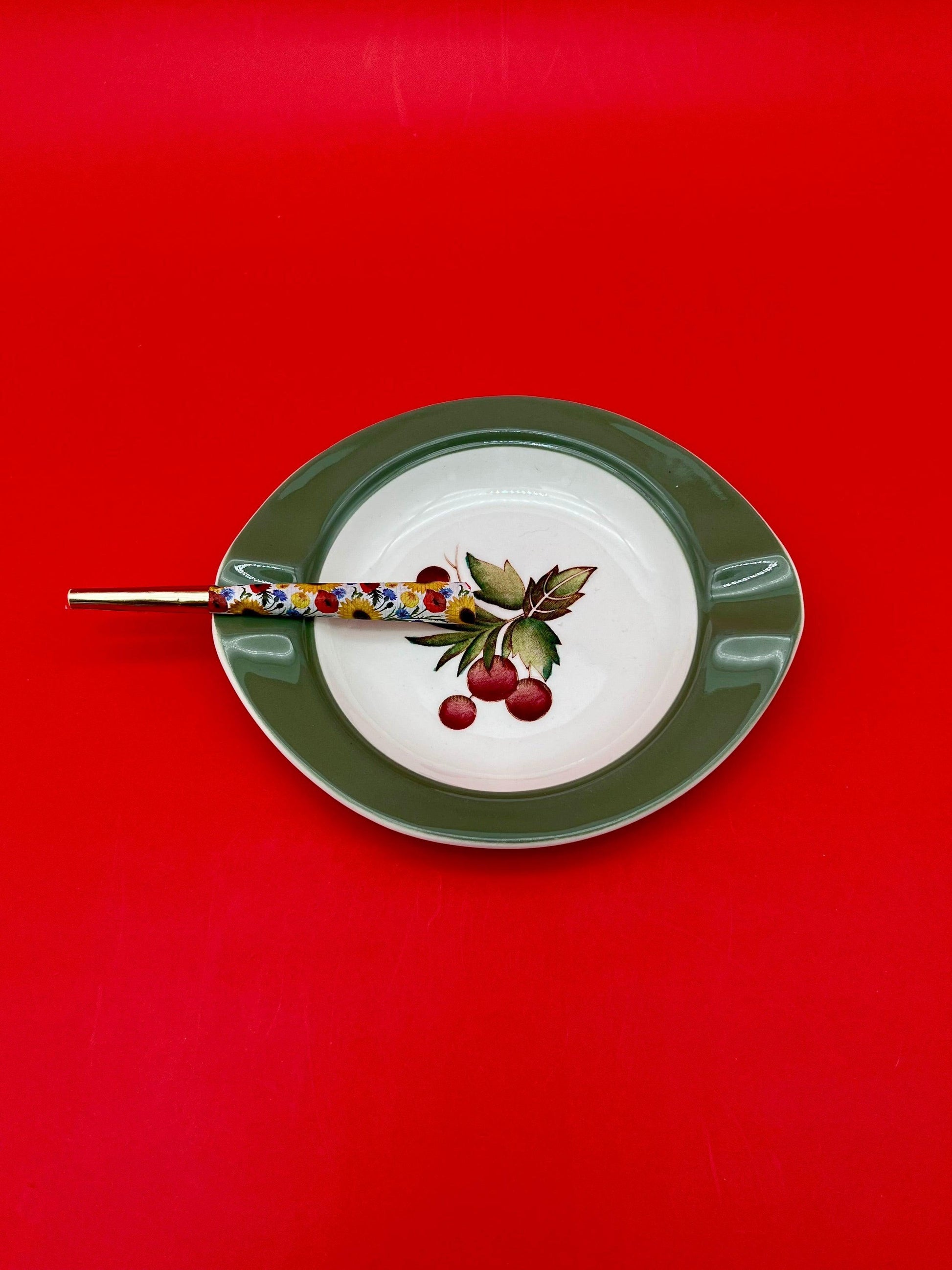 Oval white and green ceramic ashtray with a hand-painted cherry branch design, glass cigarette pick resting across it, red background.
