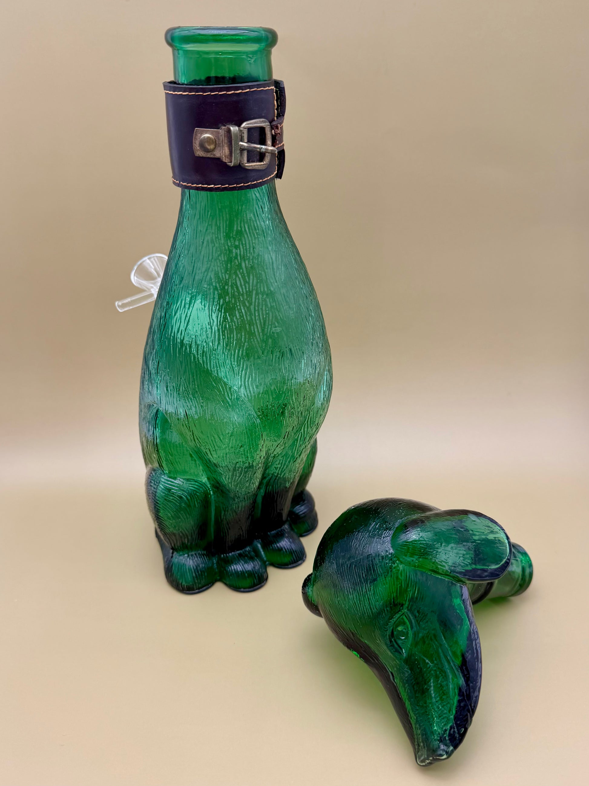 Side view of the green glass dog decanter with its separate ear-shaped glass stopper piece resting beside it, tan background.
