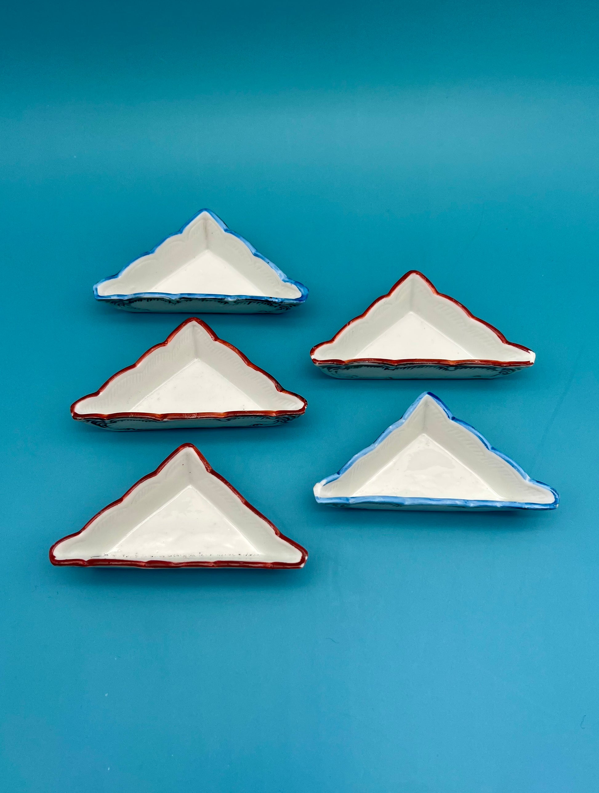 Five small triangular porcelain ashtrays with red rims from the Ludwig Set, arranged in two rows, teal background.