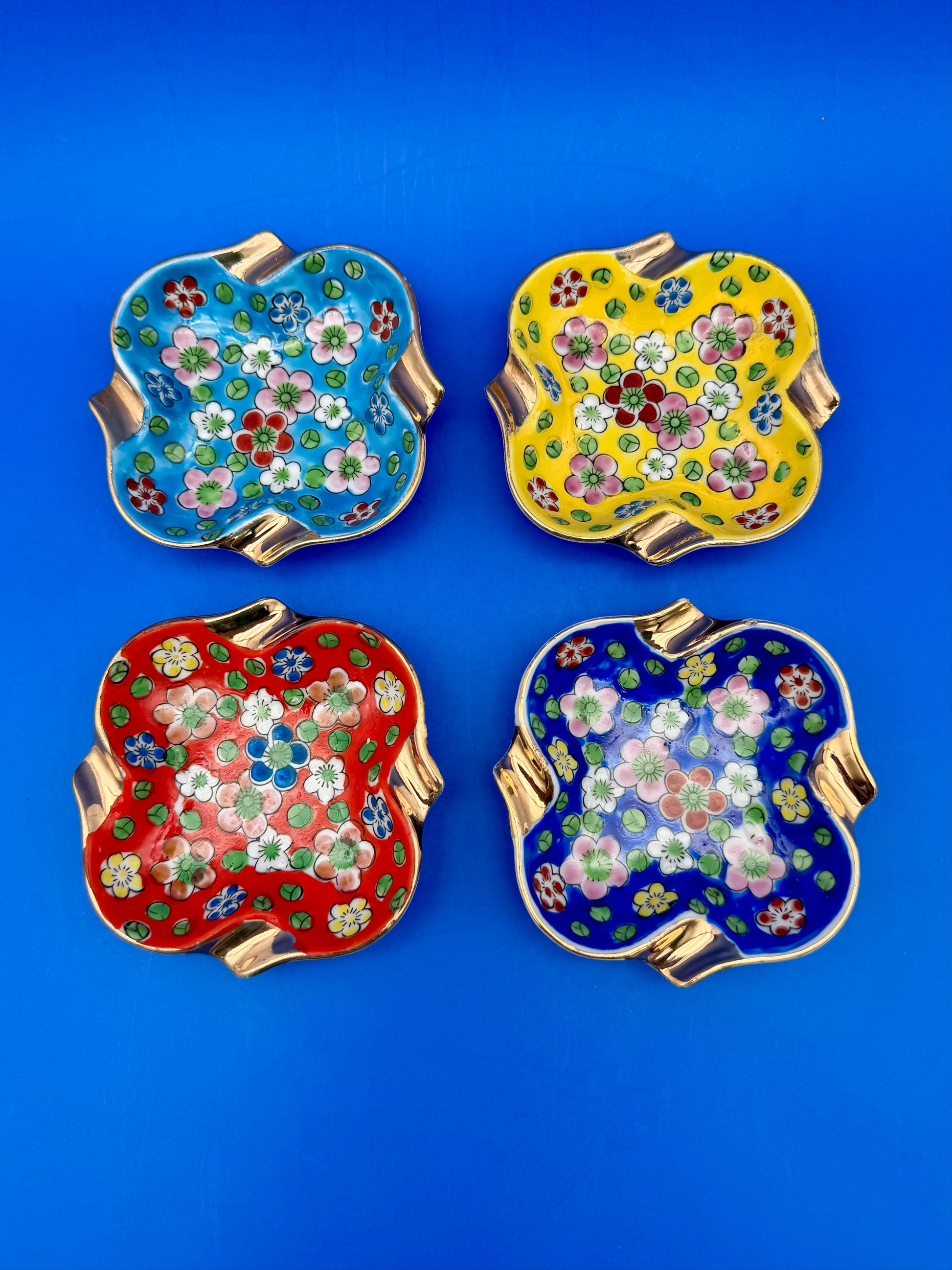 Four square ceramic ashtrays with vibrant floral patterns in teal, yellow, red, and navy blue, arranged in a grid, blue background